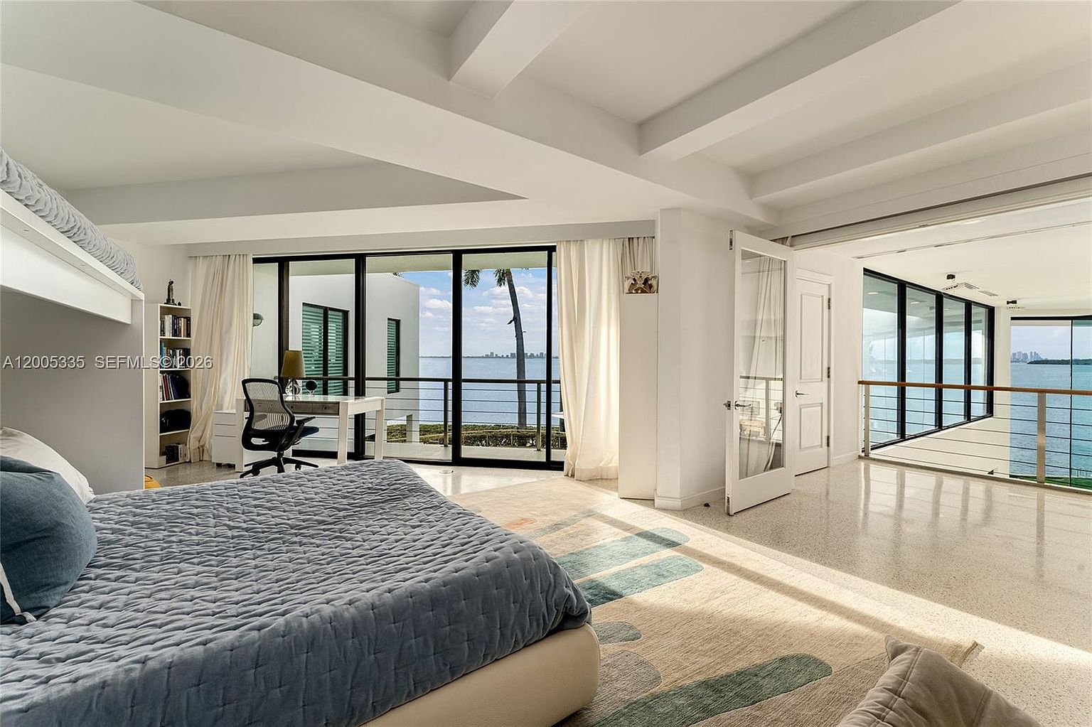 This bright and airy bedroom features a large bed with a textured blue quilt, positioned to face expansive floor-to-ceiling windows that offer a panoramic view of the water. To the side, a dedicated workspace with a desk and chair is set against the glass, while an open doorway leads to a bright, adjacent space with a glass railing overlooking the exterior. The room is finished with light-colored flooring and minimalist decor, creating a serene and open atmosphere.