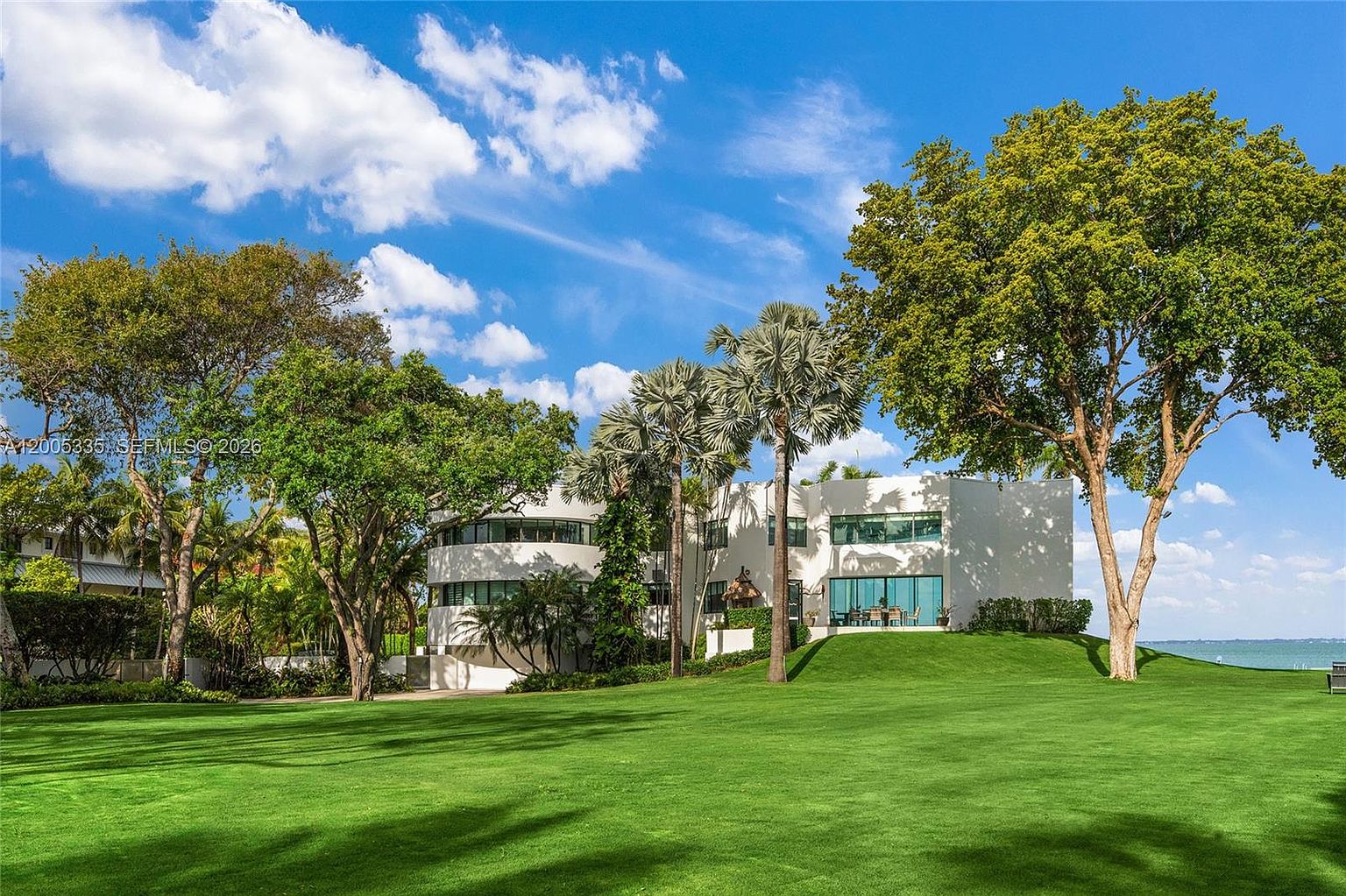 This rear view of a modern, multi-level residence showcases a distinctive curved architectural design with white exterior walls and large windows overlooking a sprawling, manicured lawn. The property is framed by mature trees and tall palm trees, creating a serene, park-like setting that extends toward the water in the distance. The perspective is captured from a low angle on the lawn, emphasizing the expansive green space and the home's unique structural silhouette.