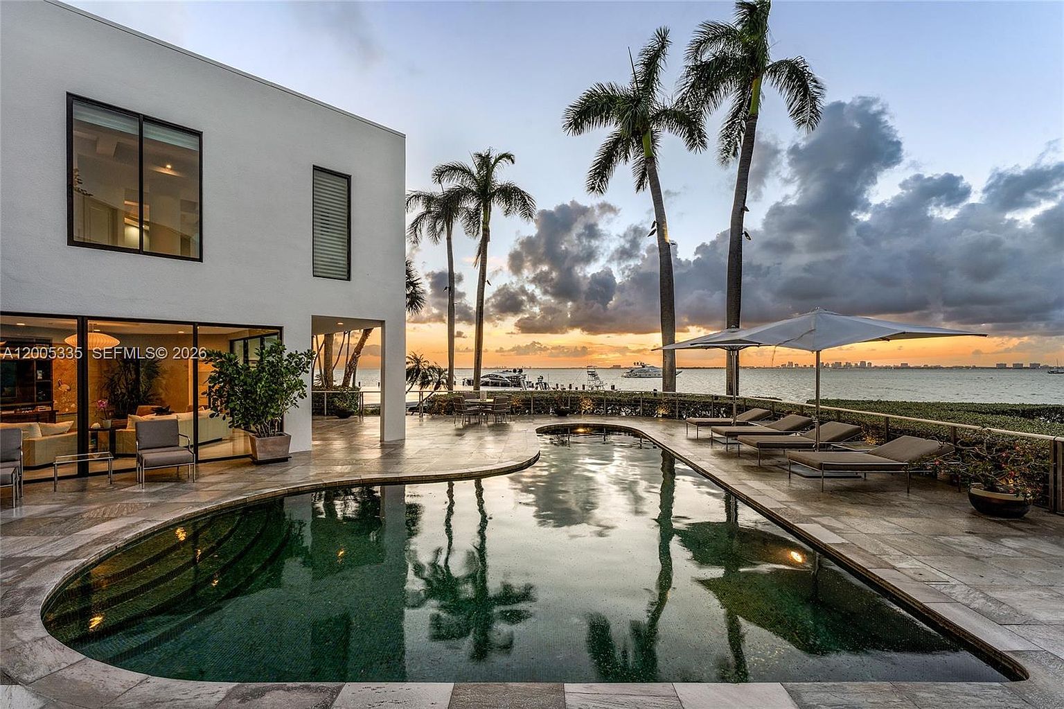 This luxurious waterfront property features a modern, white-walled home with large glass sliding doors that open onto a spacious stone-tiled patio. A curved, dark-bottomed swimming pool serves as the centerpiece, reflecting the vibrant sunset sky and the surrounding palm trees. The expansive deck includes multiple lounge chairs and an umbrella, offering a serene view of the bay and distant city skyline.