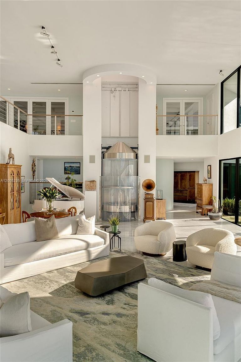 This expansive, double-height living room features a bright, open-concept design with a central glass-enclosed elevator serving as a striking architectural focal point. The space is furnished with a large white sofa, two textured cream armchairs, and a geometric coffee table, all arranged on a patterned area rug. Large windows and glass doors allow for abundant natural light, creating an airy and sophisticated atmosphere.