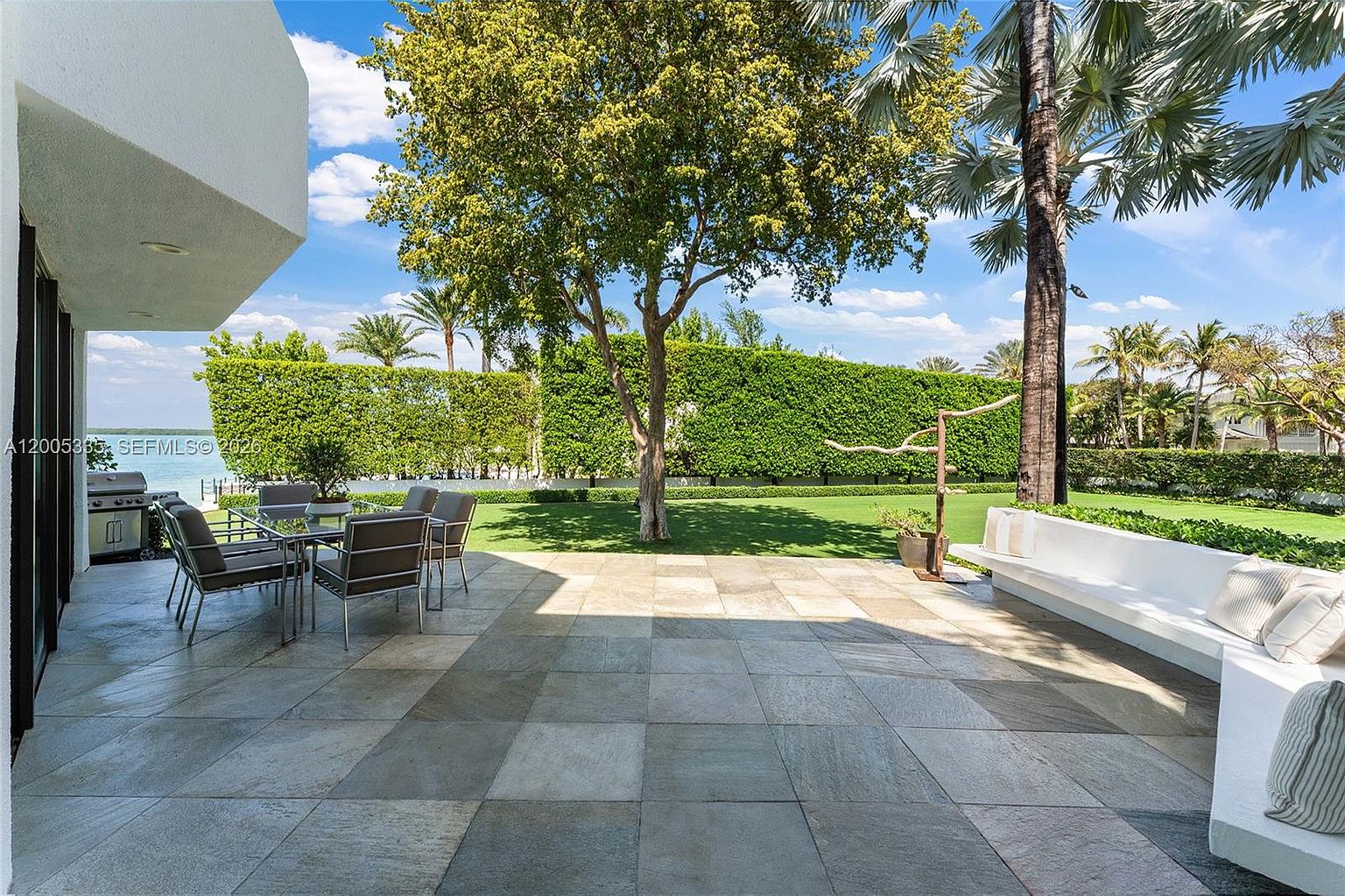 This spacious stone-paved patio offers a serene outdoor living area overlooking a lush green lawn and a waterfront view. The space is furnished with a modern dining set and a long, built-in white bench, creating an inviting atmosphere for relaxation or entertaining. Tall, manicured hedges provide privacy, while a large tree and palm trees frame the picturesque backyard setting.