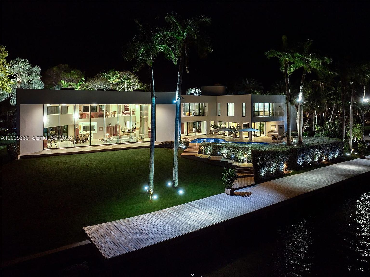 This stunning night-time aerial perspective captures a luxurious modern waterfront estate, characterized by its expansive glass walls and clean, minimalist architectural lines. The property features a sprawling manicured lawn, a private wooden dock extending into the water, and a beautifully illuminated swimming pool area nestled between the home and the shoreline. Tall palm trees and strategically placed landscape lighting enhance the sophisticated, resort-like atmosphere of this high-end residence.