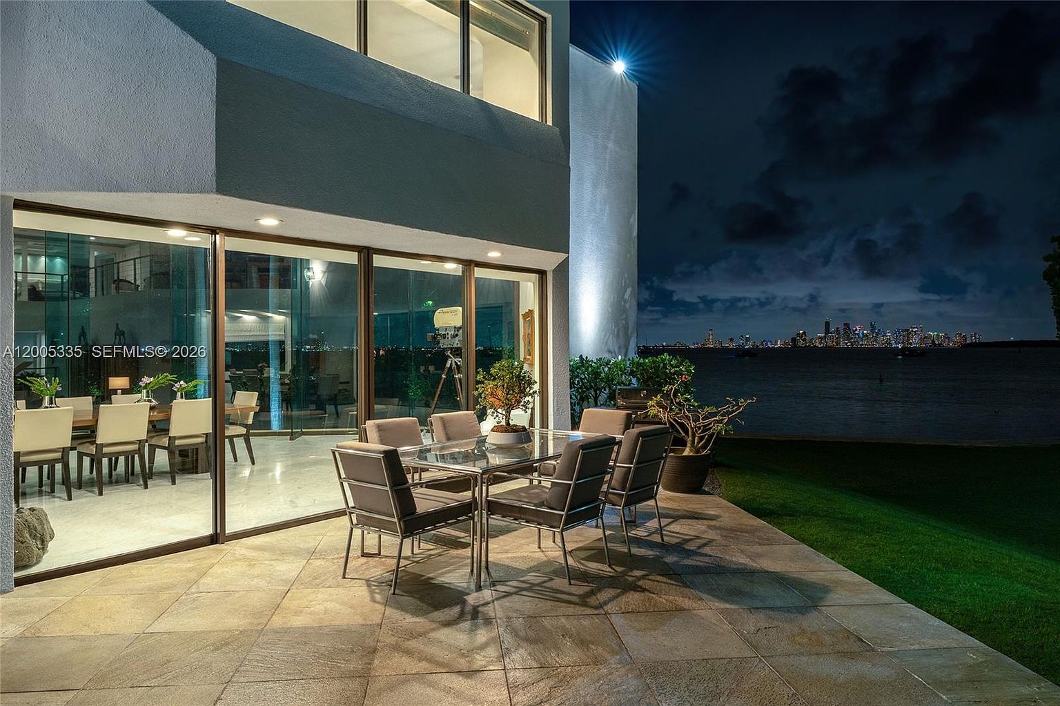 This nighttime exterior shot captures a spacious stone-tiled patio featuring a modern glass-topped dining set, perfect for outdoor entertaining. Large glass sliding doors provide a seamless transition to the interior dining area, while the expansive waterfront view showcases a distant city skyline illuminated against the night sky. The scene is framed by a manicured lawn and a contemporary architectural structure, creating a sophisticated and serene atmosphere.