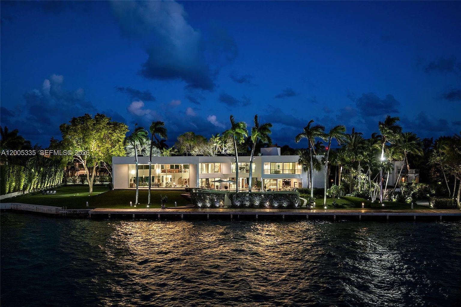 This stunning waterfront estate is captured at twilight, showcasing a contemporary architectural design with expansive glass walls that illuminate the interior. The property features a manicured lawn, mature palm trees, and a private wooden dock extending into the calm water. The cinematic perspective highlights the seamless transition between the modern residence and its serene, natural surroundings.