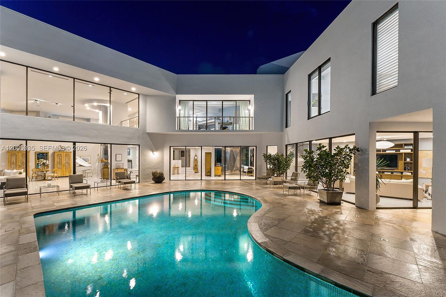 This striking contemporary residence features a U-shaped architectural design centered around a curved, illuminated swimming pool. The exterior is characterized by expansive glass walls that provide seamless indoor-outdoor connectivity, while the stone-paved patio offers ample space for lounging. Captured at twilight, the scene highlights the home's modern aesthetic and sophisticated lighting, creating an inviting and luxurious atmosphere.