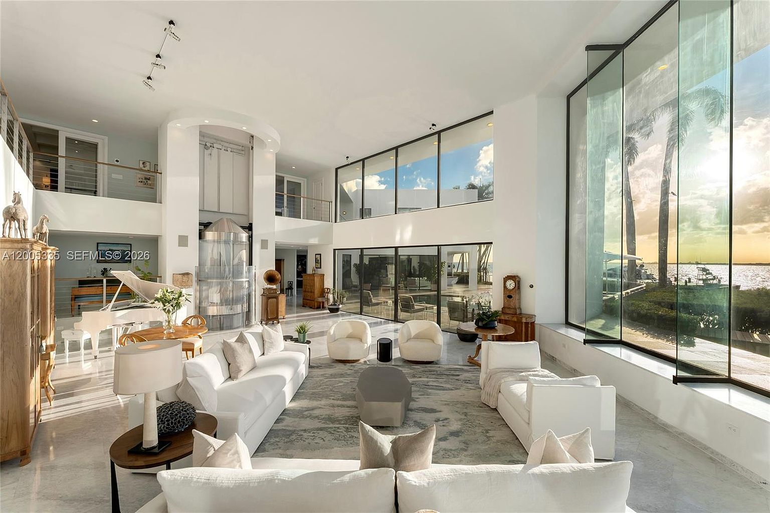 This expansive, double-height living room features a bright, open-concept design with polished light-toned flooring and a neutral color palette. A massive wall of floor-to-ceiling glass provides panoramic waterfront views, while a grand piano and contemporary white seating arrangements define the living space. The room is accented by a unique cylindrical metallic structure and a mezzanine level, creating a sophisticated and airy atmosphere.