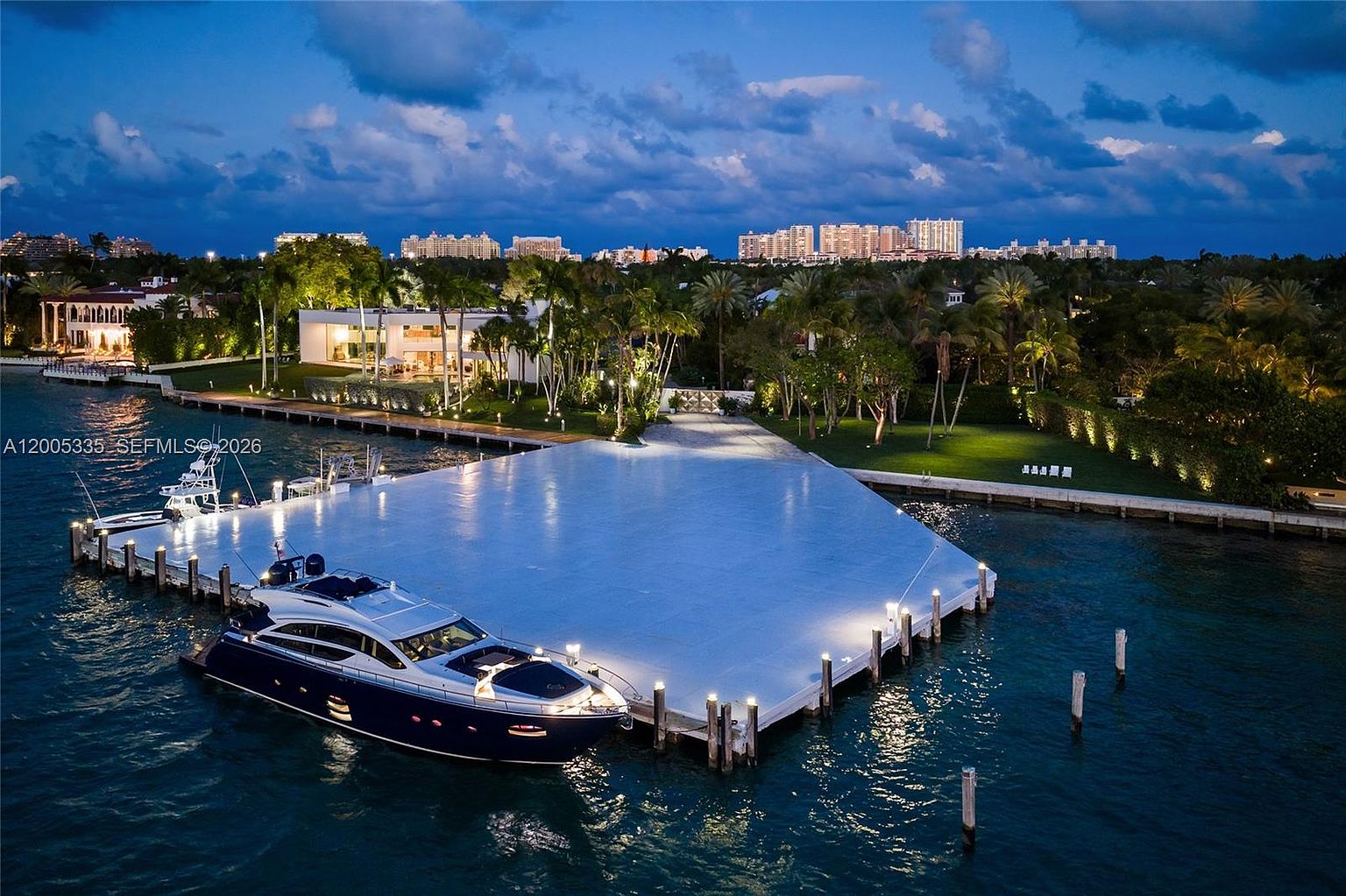 This aerial view captures a luxurious waterfront estate at twilight, featuring a large, illuminated dock extending into the water with a sleek yacht moored alongside. The property showcases a modern, expansive residence nestled among lush tropical landscaping and mature palm trees. The scene is set against a backdrop of distant city lights and a deep blue evening sky, emphasizing the property's exclusive and serene coastal atmosphere.