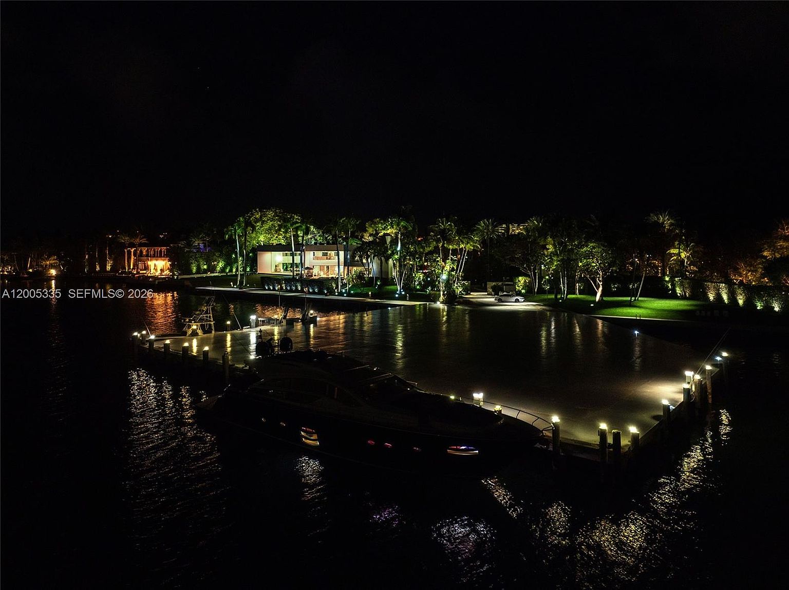 This nighttime aerial view captures a luxurious waterfront property featuring a large, illuminated private dock with a docked yacht. The scene highlights the expansive, manicured lawn and the glowing architectural lighting of the residence in the background, creating a sophisticated and serene atmosphere. The perspective emphasizes the property's prime location on the water, showcasing the seamless integration between the home and its outdoor amenities.