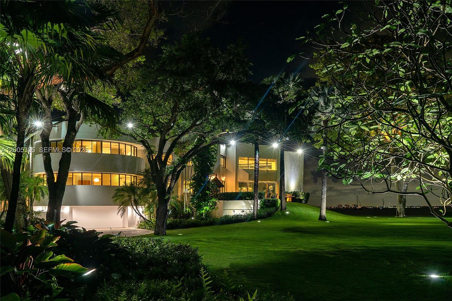 This night-time exterior view captures the rear of a modern, multi-level residence set against a lush, expansive lawn. The home features a distinctive curved architectural design with large, horizontal windows that glow warmly, contrasting with the dark, manicured landscape. Tall, mature trees frame the structure, while the perspective highlights the property's connection to the waterfront in the distance.