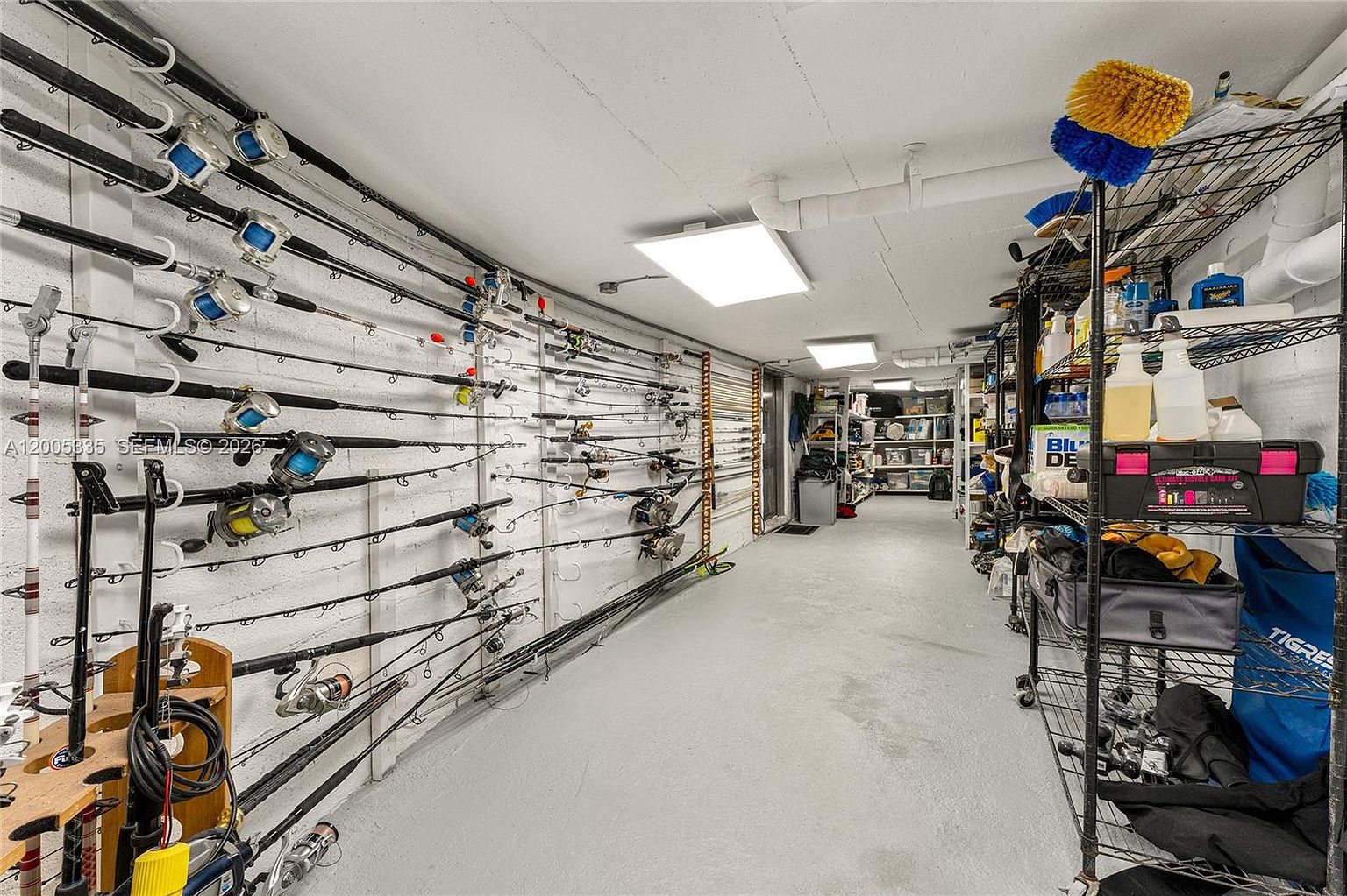 This utilitarian storage space features a long, narrow layout with white-painted masonry walls lined with numerous fishing rods mounted on racks. To the right, a metal shelving unit holds various cleaning supplies and gear, while the concrete floor leads toward a deeper storage area in the background. The space is illuminated by overhead panel lighting, creating a functional and organized environment for hobbyist storage.