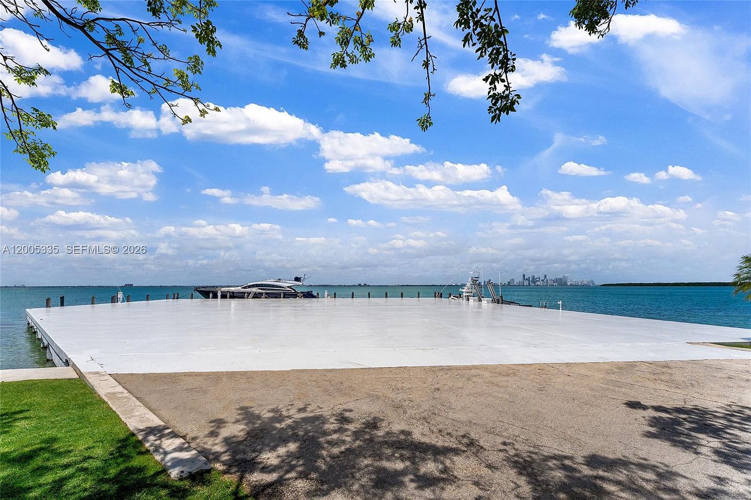 This image captures a expansive, flat concrete dock or pier extending out into a calm, blue bay under a bright, partly cloudy sky. A sleek, modern yacht is moored alongside the structure, which provides a clear, unobstructed view of the distant city skyline across the water. The perspective is from the shore looking out, emphasizing the generous outdoor space and the prime waterfront location.
