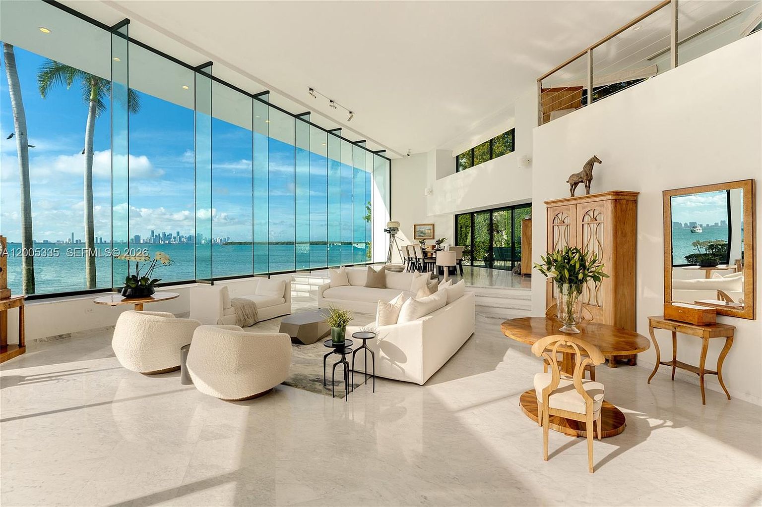 This expansive, light-filled living room features dramatic floor-to-ceiling windows that provide an unobstructed, panoramic view of the ocean and distant city skyline. The space is anchored by a large, neutral-toned sectional sofa and curved accent chairs, all set upon a polished stone floor. A tall wooden cabinet topped with a horse sculpture and a nearby dining area add a touch of elegance to the open-concept layout.