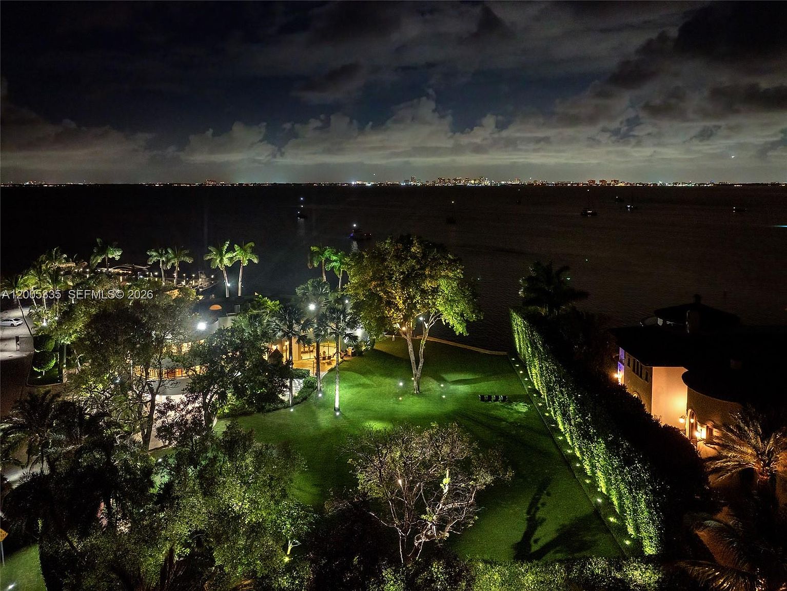 This nighttime aerial view captures a sprawling waterfront estate featuring a lush, manicured lawn illuminated by landscape lighting. The property includes mature trees, a private dock area, and a large residence, all set against the backdrop of a calm bay and a distant city skyline. The perspective provides a cinematic overview of the expansive grounds and the serene, high-end coastal setting.