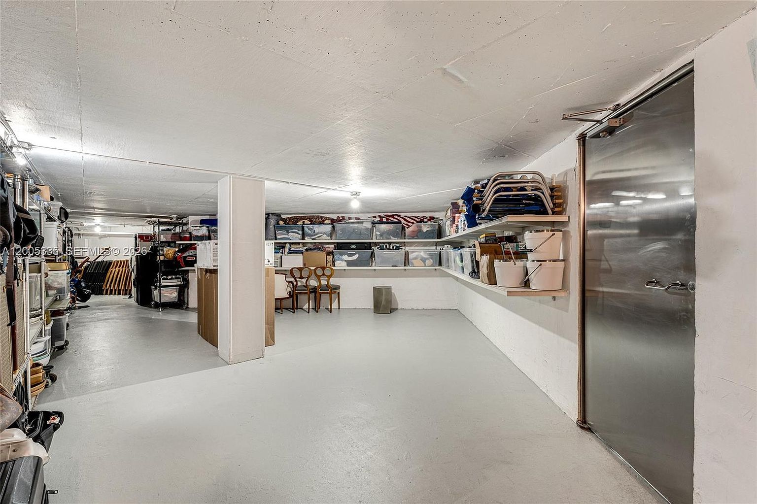 This image shows a spacious, utilitarian basement storage area with a concrete floor and white-painted walls. The room features extensive shelving units along the walls, filled with various storage bins and household items, and a heavy-duty metal door on the right. The perspective is from the center of the room, looking toward the back corner, highlighting the ample storage capacity and functional layout of the space.