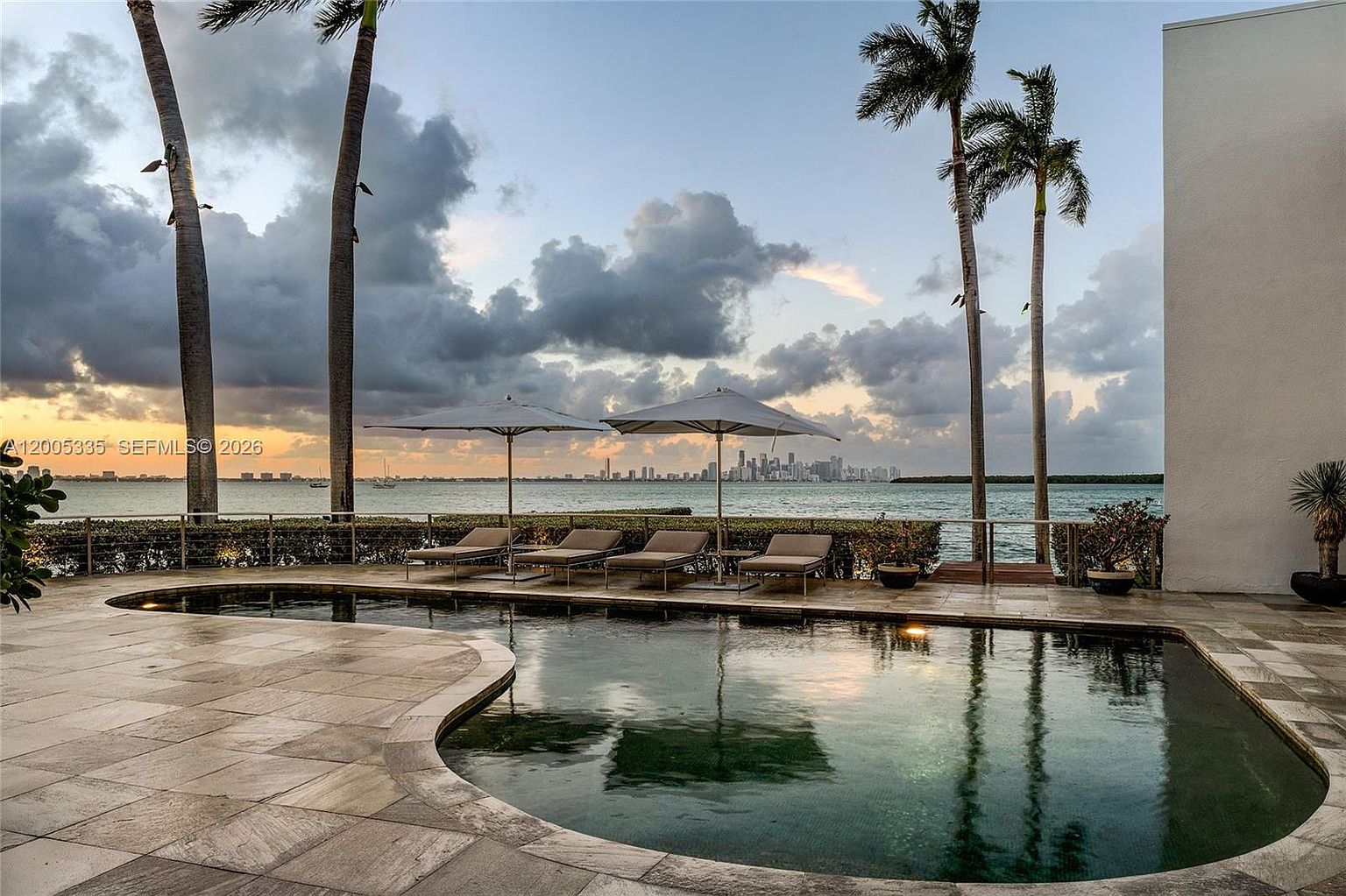 This serene waterfront pool area features a curved, dark-bottomed pool set against a backdrop of the ocean and a distant city skyline. The patio is paved with light-toned stone and includes a row of lounge chairs shaded by umbrellas, framed by tall palm trees. The perspective is a low-angle shot looking out toward the water, emphasizing the tranquil and luxurious outdoor living space.