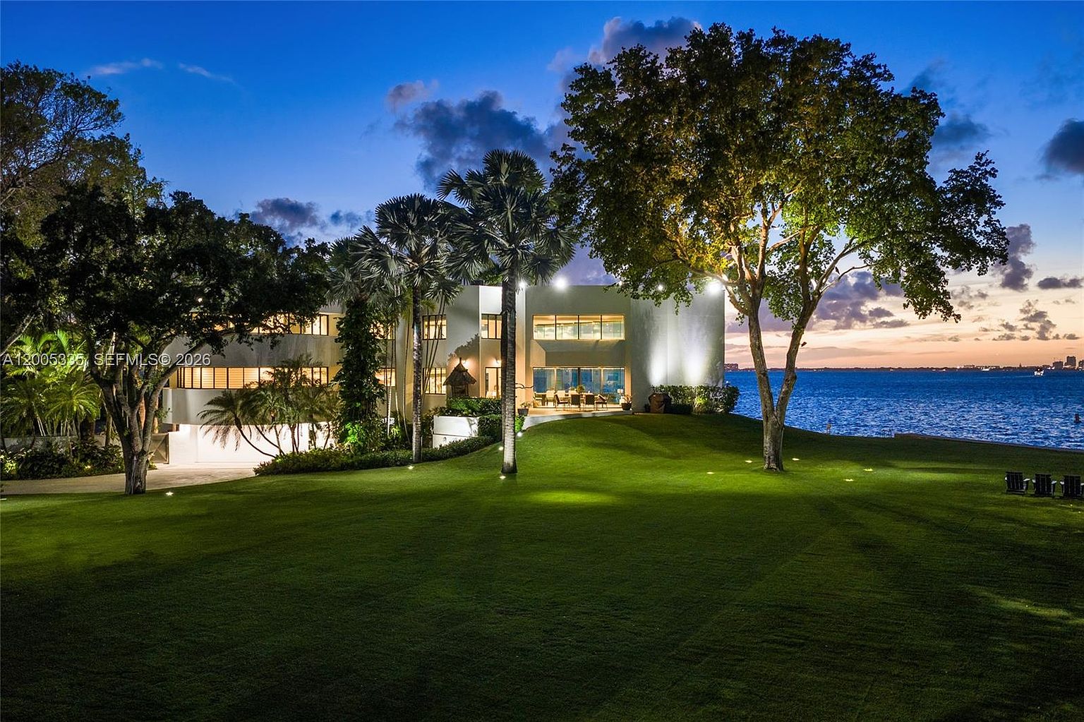 This stunning waterfront estate is captured at twilight, showcasing a modern architectural design with expansive glass windows that overlook a vast, manicured lawn. The property features mature trees and palm trees that frame the structure, while the calm water of the bay stretches out toward the horizon under a soft, colorful sunset sky. The scene is illuminated by warm exterior lighting, creating an inviting and luxurious atmosphere.