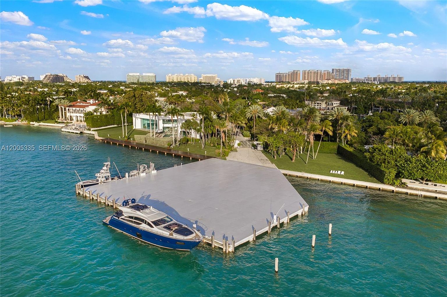 This high-angle aerial view captures a luxurious waterfront property featuring a large, modern white residence situated on a lush, palm-lined lot. A prominent, expansive gray dock extends into the turquoise water, where a sleek blue and white yacht is moored. The surrounding landscape is meticulously manicured, offering a serene and upscale coastal atmosphere with a distant cityscape visible on the horizon.