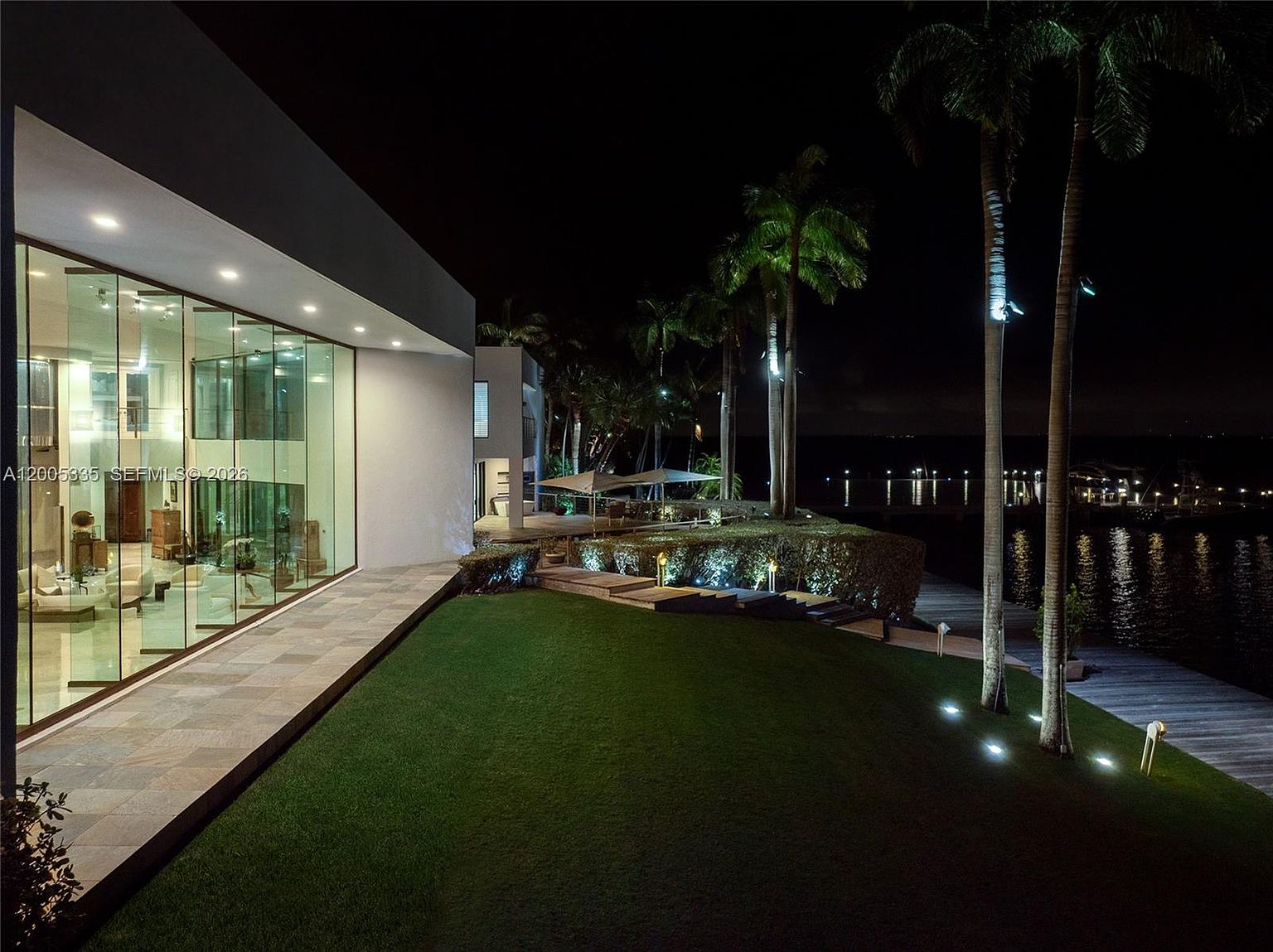 This night-time view captures the rear of a modern luxury residence, characterized by expansive floor-to-ceiling glass walls that reveal a brightly lit interior. The property features a manicured lawn, stone-paved pathways, and tall palm trees illuminated by landscape lighting, leading toward a private wooden dock over the water. The cinematic perspective emphasizes the seamless transition between the sleek architectural design and the serene waterfront setting.