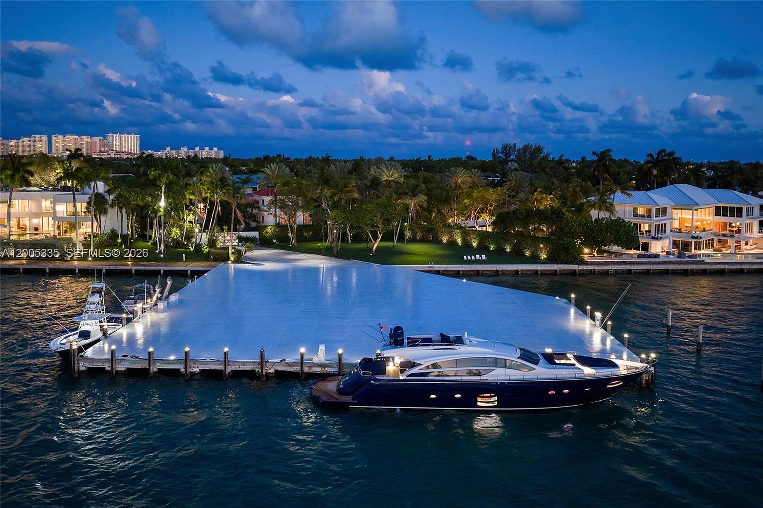 This aerial perspective captures a luxurious waterfront property at dusk, featuring a large, illuminated private dock extending into the calm water. A sleek, dark blue yacht is moored alongside the dock, while the surrounding landscape showcases lush tropical greenery and modern residential architecture with warm interior lighting. The scene conveys an atmosphere of exclusivity and high-end coastal living.