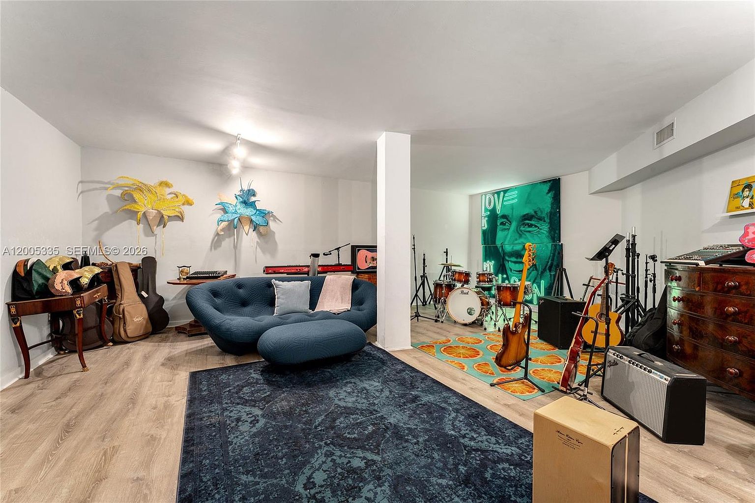 This spacious, multi-functional basement area is currently set up as a creative music studio and lounge. The room features light-toned wood-look flooring, a distinctive blue curved sofa with a matching ottoman, and a vibrant area rug with a citrus-slice pattern beneath a drum kit and musical instruments. A large, bold portrait of Bob Marley serves as a focal point on the back wall, adding a personalized artistic touch to this versatile living space.