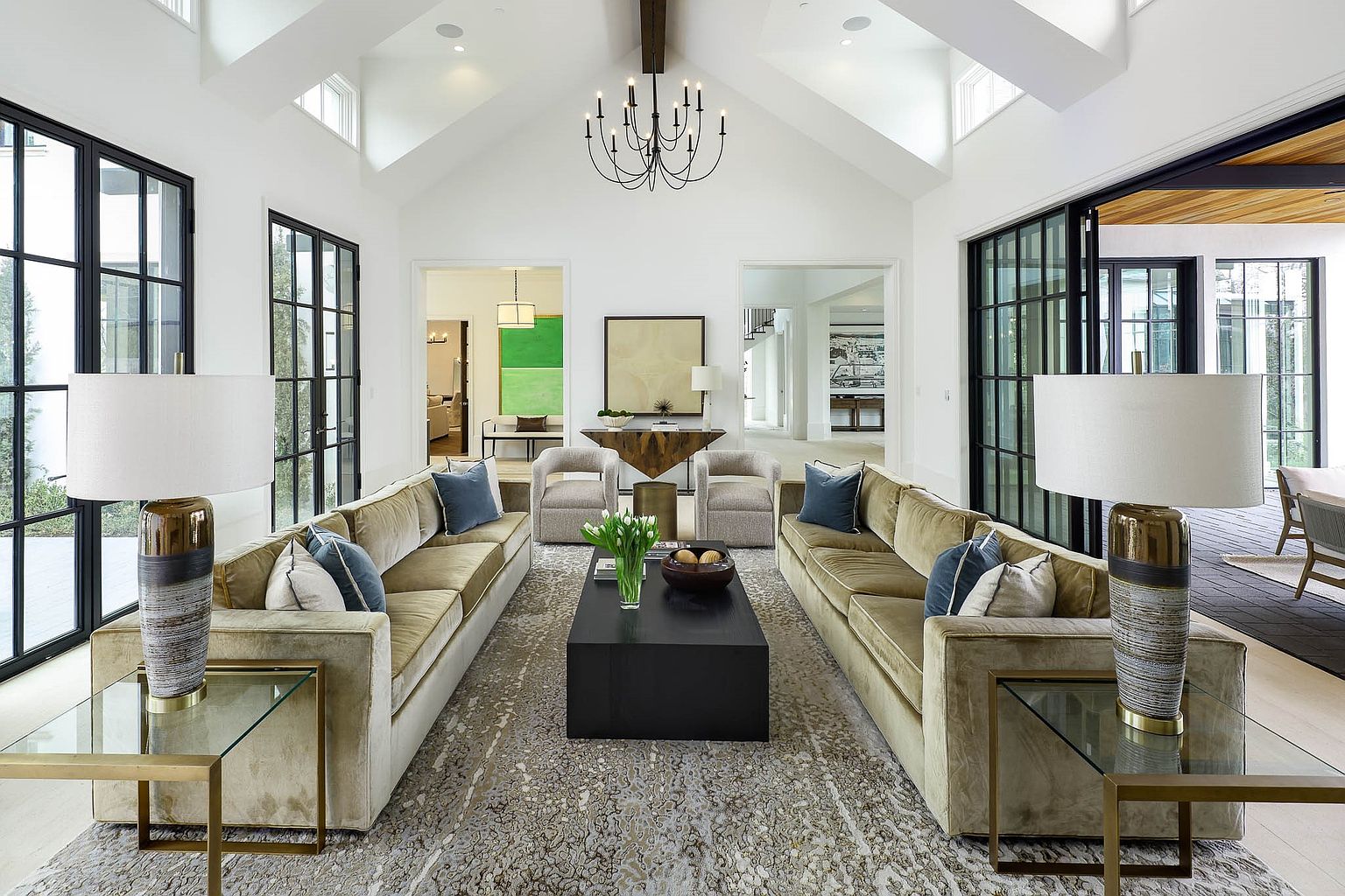 This elegant, symmetrical living room features a vaulted ceiling with exposed beams and a central chandelier, creating a grand and airy atmosphere. Two plush, velvet-upholstered sofas face each other across a dark rectangular coffee table, anchored by a large, textured area rug. Large, black-framed windows flank the space, providing abundant natural light and views of the surrounding outdoor areas.