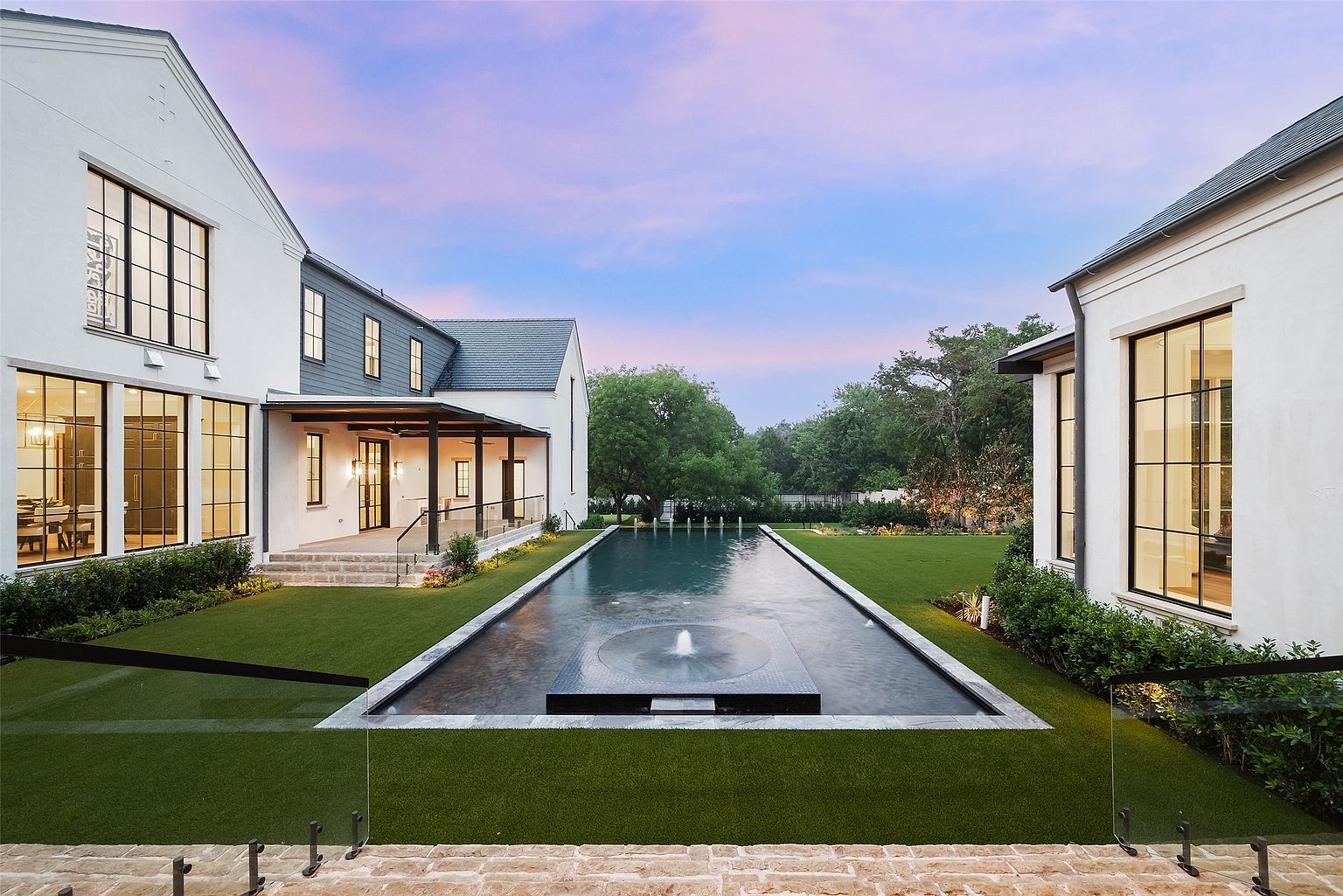 This stunning rear exterior view showcases a modern luxury residence featuring a long, rectangular lap pool as the central focal point. The home is characterized by white stucco walls, dark-framed windows, and a covered patio area, all set against a backdrop of a manicured lawn and mature trees under a soft twilight sky. The perspective is centered, emphasizing the symmetrical design and the elegant transition between the indoor living spaces and the outdoor recreational area.