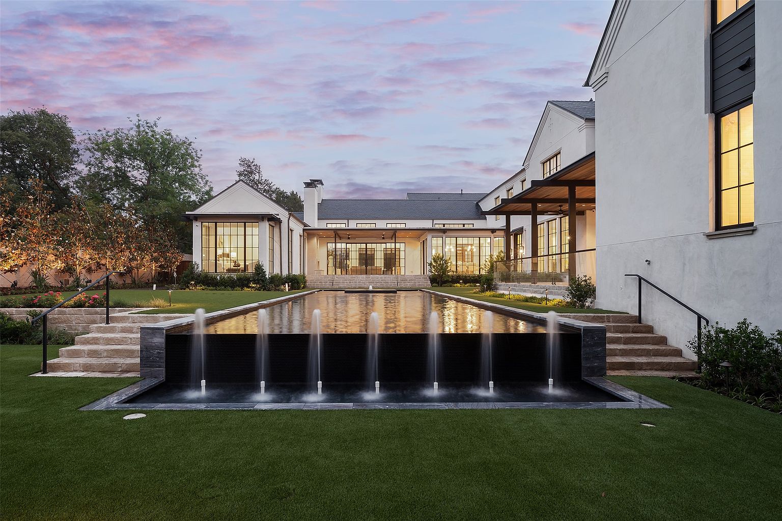 This stunning rear exterior view showcases a luxurious modern estate featuring a long, rectangular infinity-edge pool with integrated water fountains. The home is characterized by white stucco walls, large windows with dark frames, and a covered patio area that connects the indoor and outdoor living spaces. Manicured green lawns and stone staircases frame the pool, creating a sophisticated and serene atmosphere at dusk.