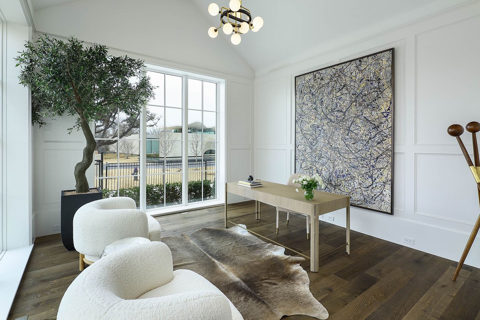 This sophisticated home office features a minimalist light wood desk positioned in front of a large, abstract expressionist painting. The room is anchored by a natural hide rug on dark wood flooring and includes two plush, cream-colored curved chairs near a large window that provides ample natural light. A modern, multi-globe chandelier hangs from the vaulted ceiling, adding a contemporary touch to the bright, white-walled space.
