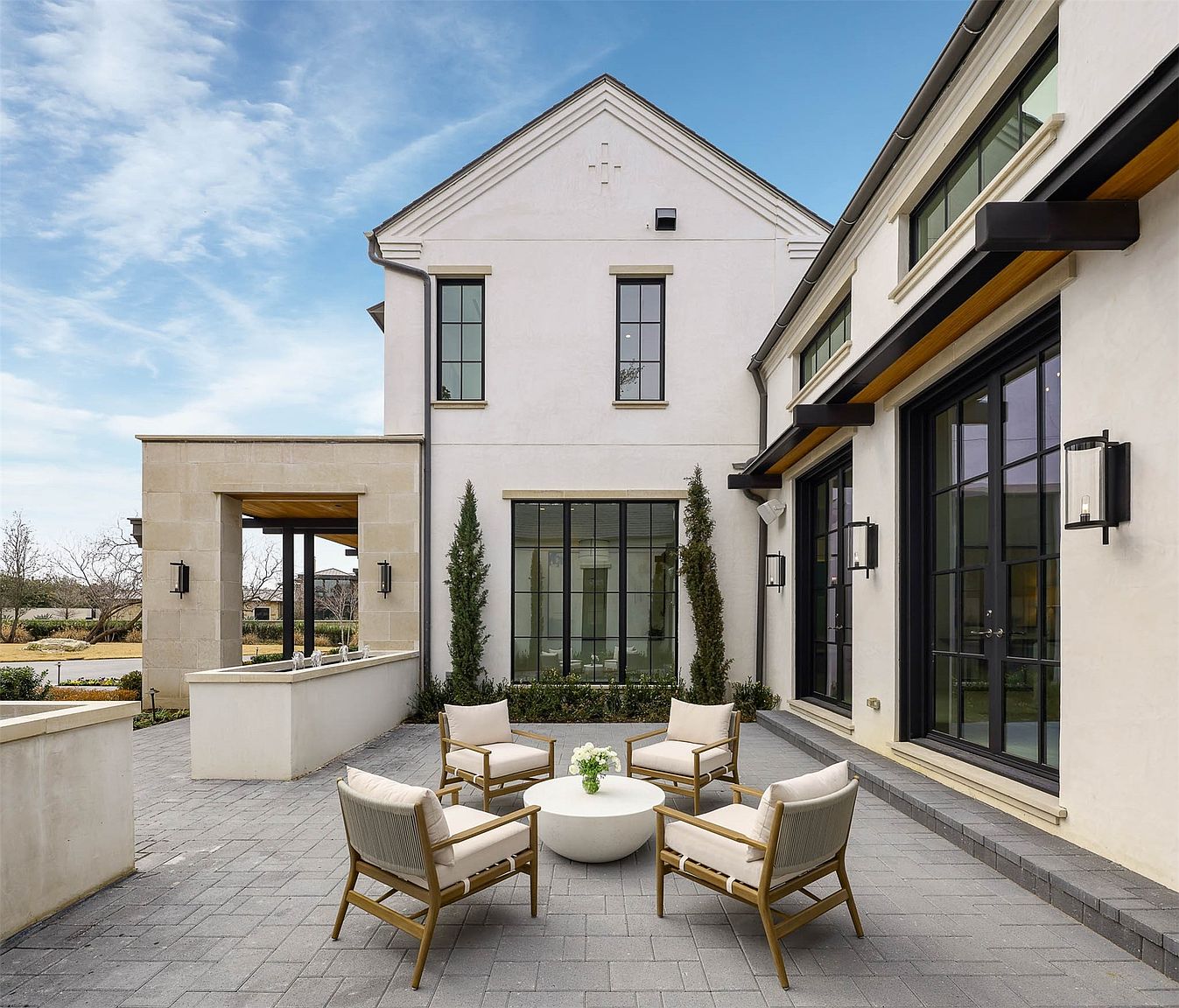This elegant outdoor patio features a clean, modern aesthetic with light-colored stucco walls and dark-framed windows. The space is furnished with four wooden armchairs arranged around a circular white coffee table, set against a backdrop of manicured greenery and a stone-clad water feature. The paved stone flooring and symmetrical architectural lines create a sophisticated and inviting atmosphere for outdoor relaxation.