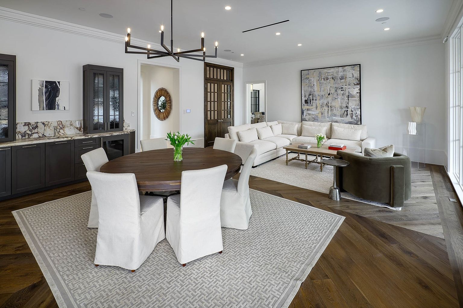 This open-concept living and dining area features a sophisticated, neutral color palette with dark wood flooring and a geometric patterned area rug. The space includes a round wooden dining table surrounded by slipcovered chairs, a built-in wet bar with dark cabinetry, and a plush sectional sofa arranged for comfortable seating. Large windows along the right wall provide ample natural light, creating an airy and inviting atmosphere suitable for both entertaining and relaxation.