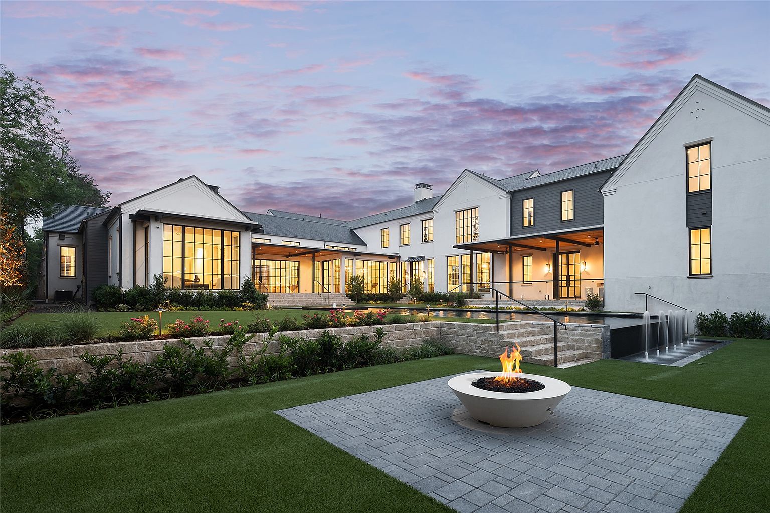 This expansive rear exterior view showcases a luxurious modern farmhouse estate at dusk, featuring a multi-level layout with white stucco and dark siding accents. The property includes a large swimming pool with integrated water features, a stone-paved patio area with a circular fire bowl, and meticulously manicured landscaping. Large windows and glass doors create a seamless connection between the interior living spaces and the outdoor entertainment areas.