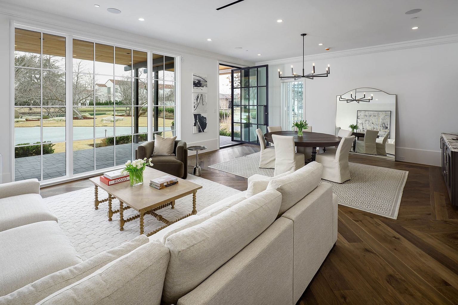 This open-concept living and dining area features rich, dark wood flooring arranged in a herringbone pattern and bright, neutral-toned furnishings. Large windows provide an expansive view of the outdoor landscape, while a modern chandelier hangs above the circular dining table. The space is designed with a sophisticated, airy aesthetic, highlighted by a large floor mirror and minimalist wall art.