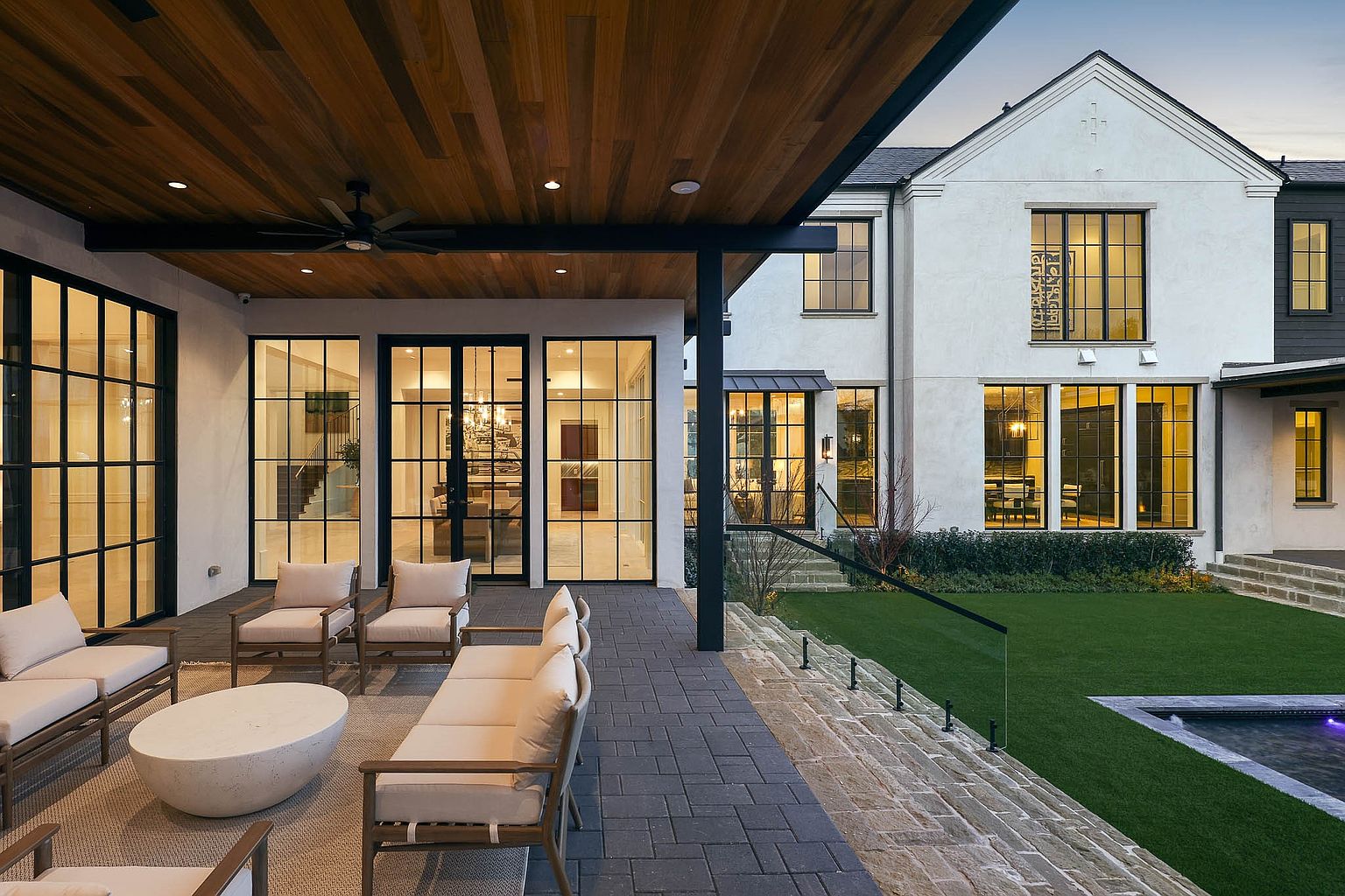 This elegant covered patio features a warm wood-paneled ceiling with a built-in fan and recessed lighting, overlooking a manicured lawn and a swimming pool. The space is furnished with comfortable outdoor seating, including armchairs and a sofa arranged on a rug, set against a backdrop of large, grid-paned windows and doors that connect the indoor and outdoor living areas. The architectural style is sophisticated and modern, emphasizing a seamless transition between the interior and the exterior landscape.