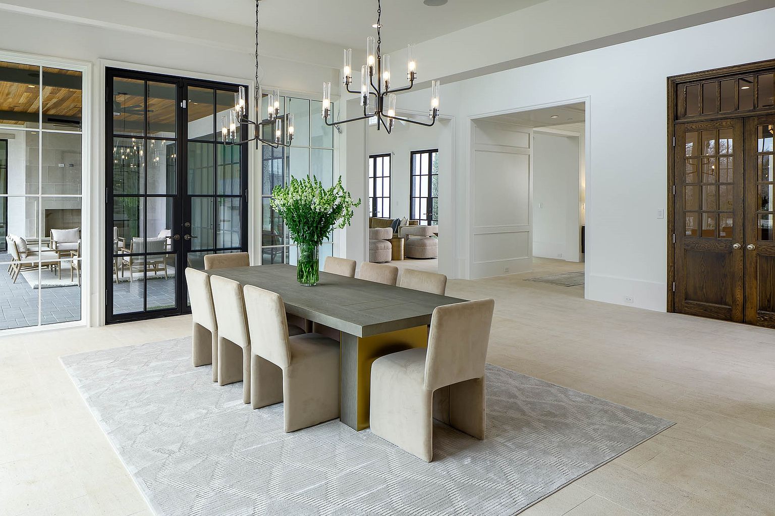 This elegant dining room features a large rectangular table with a grey top and a gold-toned base, surrounded by eight upholstered chairs. The space is illuminated by two modern, multi-armed chandeliers and is anchored by a large, light-colored patterned rug. Large glass-paned doors provide a view into an outdoor patio area, while a set of dark wood double doors adds a rich contrast to the bright, neutral interior.