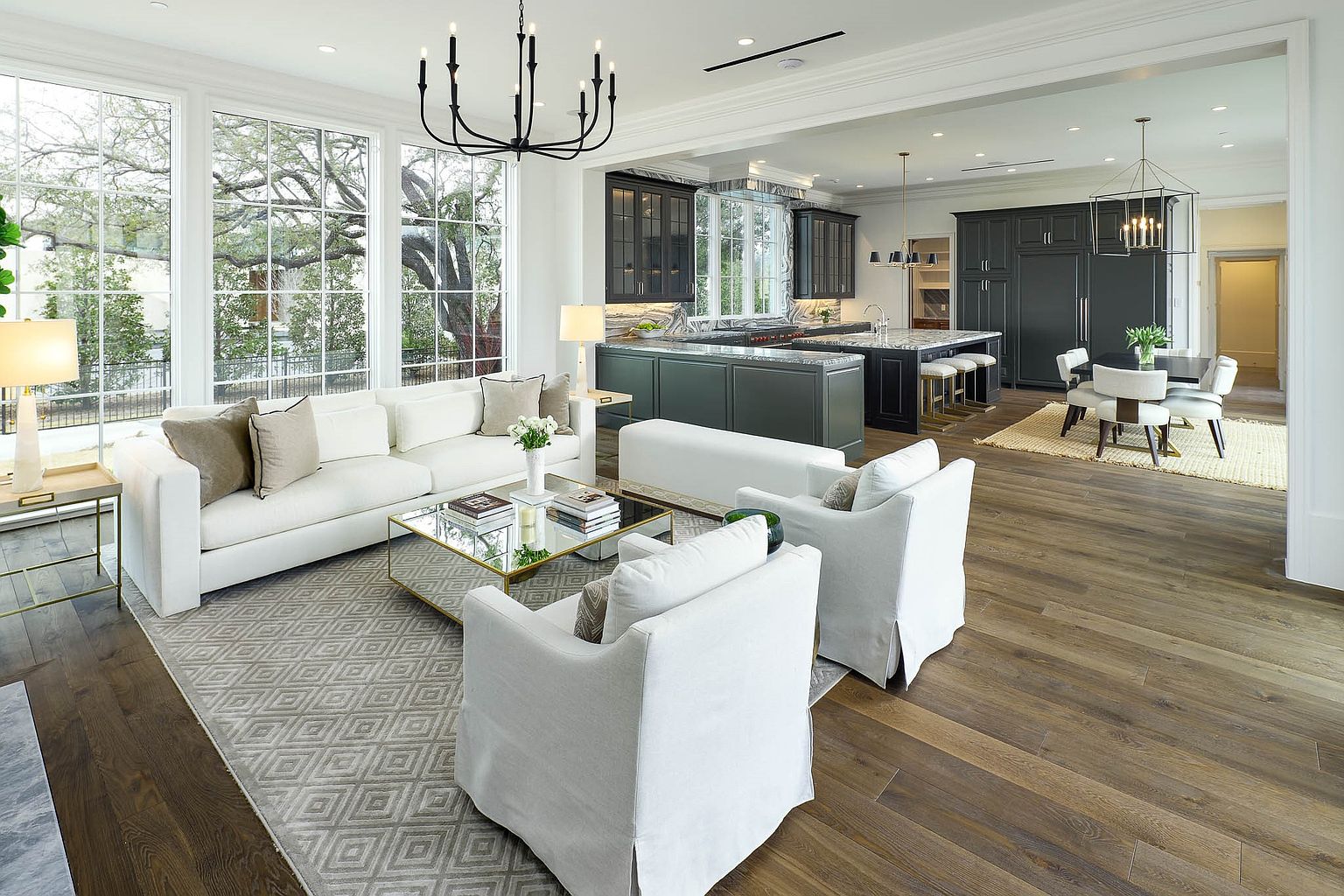 This open-concept living space features a bright, neutral color palette with a plush white sofa and matching armchairs arranged around a glass-topped coffee table. Large windows along the left wall provide abundant natural light, while the adjacent kitchen and dining area showcase dark cabinetry and modern light fixtures. The room is anchored by a patterned area rug over warm-toned wood flooring, creating a sophisticated and airy atmosphere suitable for entertaining.