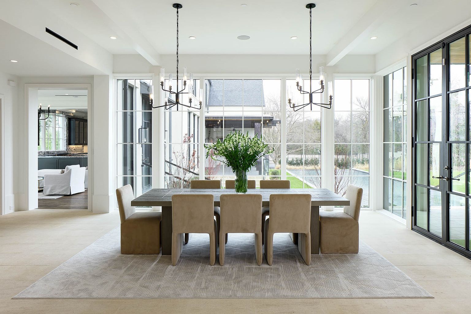 This elegant dining room features a long, dark-toned table surrounded by eight upholstered chairs in a neutral beige fabric. The space is defined by expansive, floor-to-ceiling windows that offer a clear view of the outdoor landscape and pool area, flooding the room with natural light. Two black metal chandeliers hang above the table, adding a sophisticated, modern touch to the bright and airy interior.