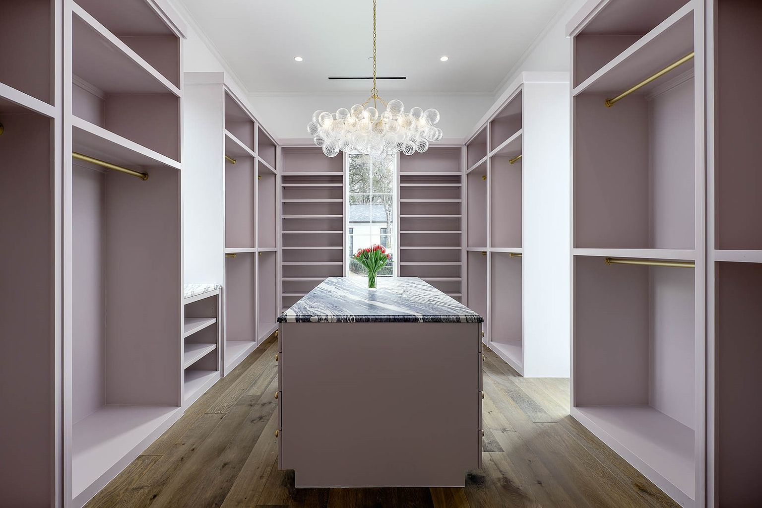 This spacious walk-in closet features custom light-toned cabinetry with integrated brass hanging rods and open shelving. A central island with a dark, patterned stone countertop serves as a focal point, topped with a small vase of red tulips. The room is illuminated by a decorative glass bubble chandelier, and the perspective looks directly down the center aisle toward a window that provides natural light.