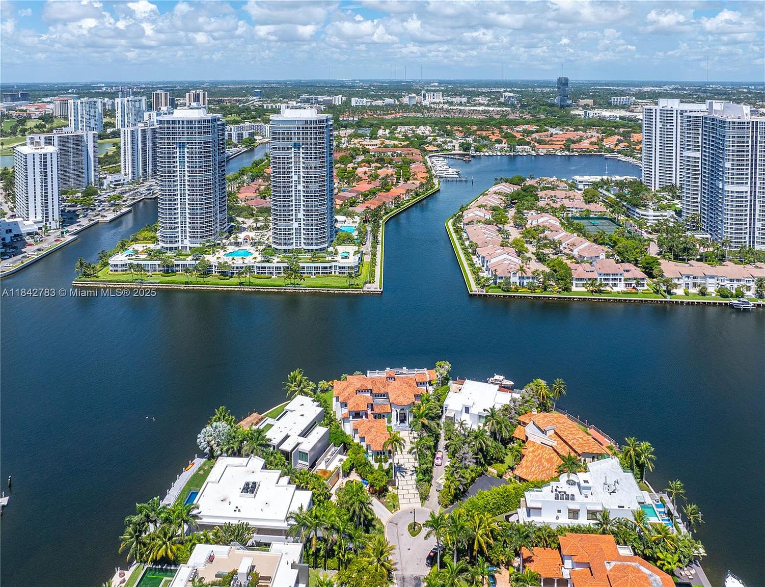 This high-angle aerial perspective captures a luxurious waterfront residential enclave characterized by a complex network of canals and waterways. In the foreground, a sprawling estate with a terracotta-tiled roof and lush tropical landscaping sits on a point, while the background features towering high-rise residential buildings and dense suburban development. The scene conveys a sense of prestige and coastal living, highlighting the prime location and expansive water views.