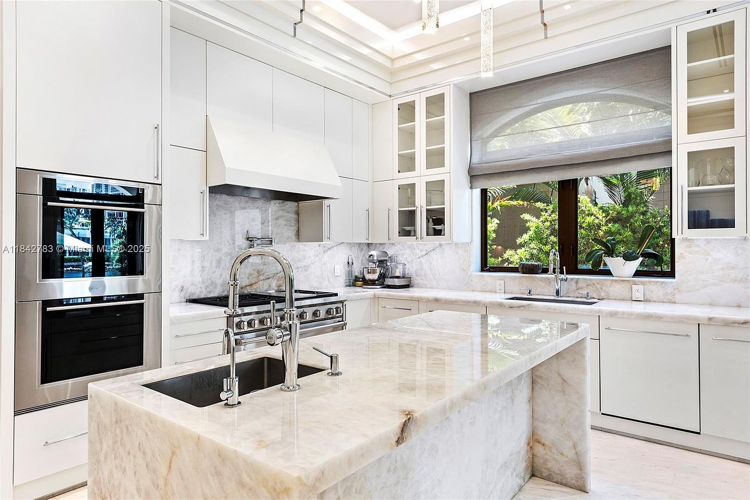 This bright, high-end kitchen features extensive stone surfaces on the countertops, backsplash, and the large central island. The space is equipped with professional-grade stainless steel appliances, including a double wall oven and a gas range, complemented by crisp white cabinetry and glass-fronted upper cabinets. A large window above the secondary sink provides a view of lush greenery, while recessed lighting and elegant fixtures create a sophisticated and airy atmosphere.