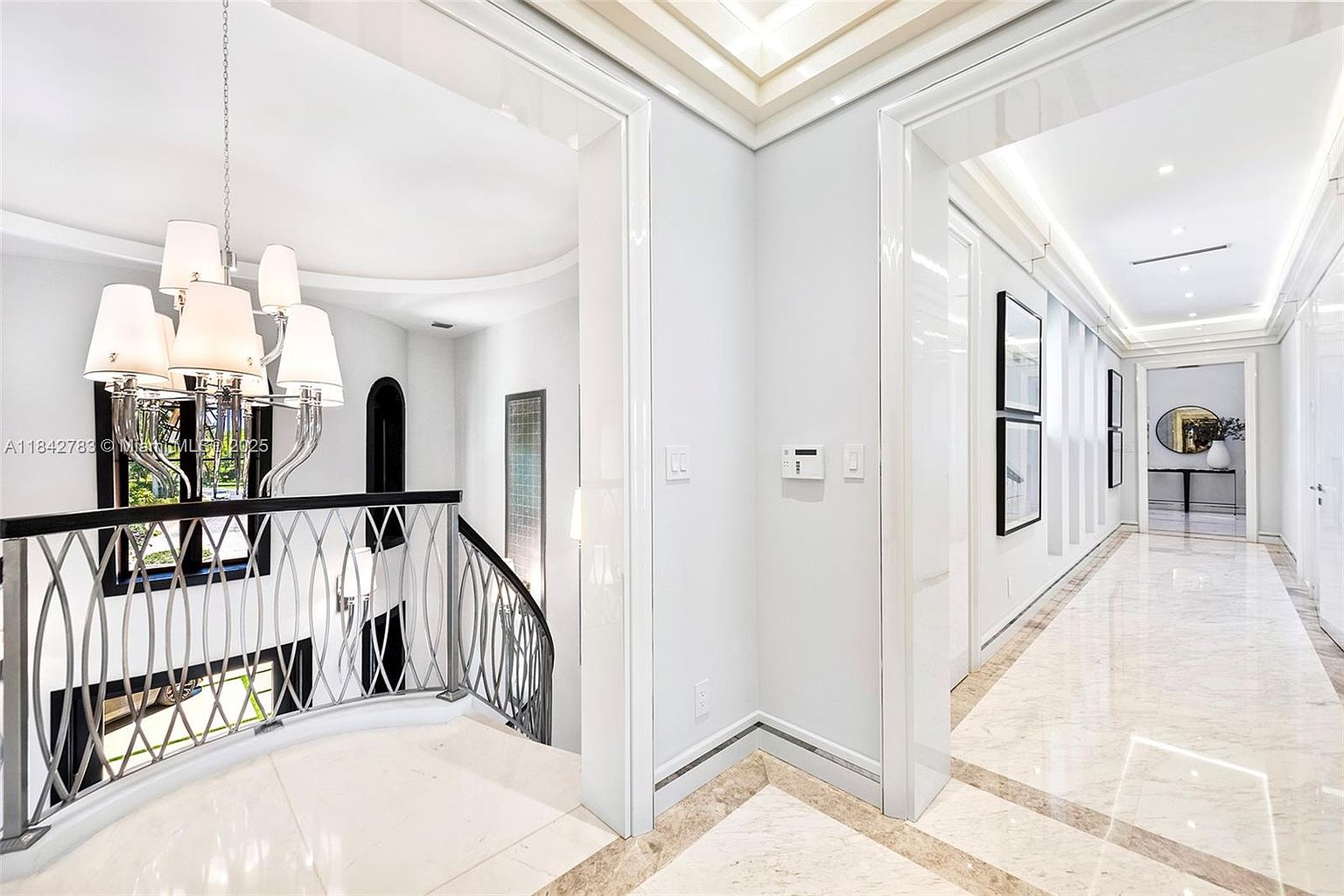 This elegant interior view captures a grand landing area featuring a sophisticated chandelier and a decorative metal railing overlooking the level below. A long, bright hallway extends to the right, characterized by polished light-colored stone flooring and recessed ceiling lighting. The space exudes a luxurious, modern aesthetic with clean lines and a neutral color palette.