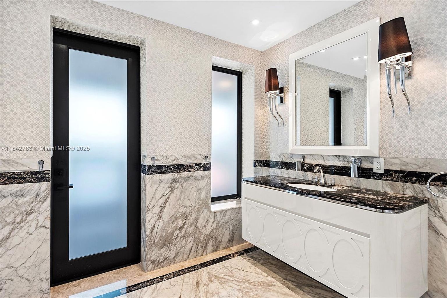 This elegant bathroom features a floating white vanity topped with a dark, veined stone countertop and a single undermount sink. The walls are adorned with a combination of patterned wallpaper and stone wainscoting, complemented by a matching stone floor border. A large mirror is flanked by two modern wall sconces, while a frosted glass door and a tall, narrow window provide natural light and privacy.