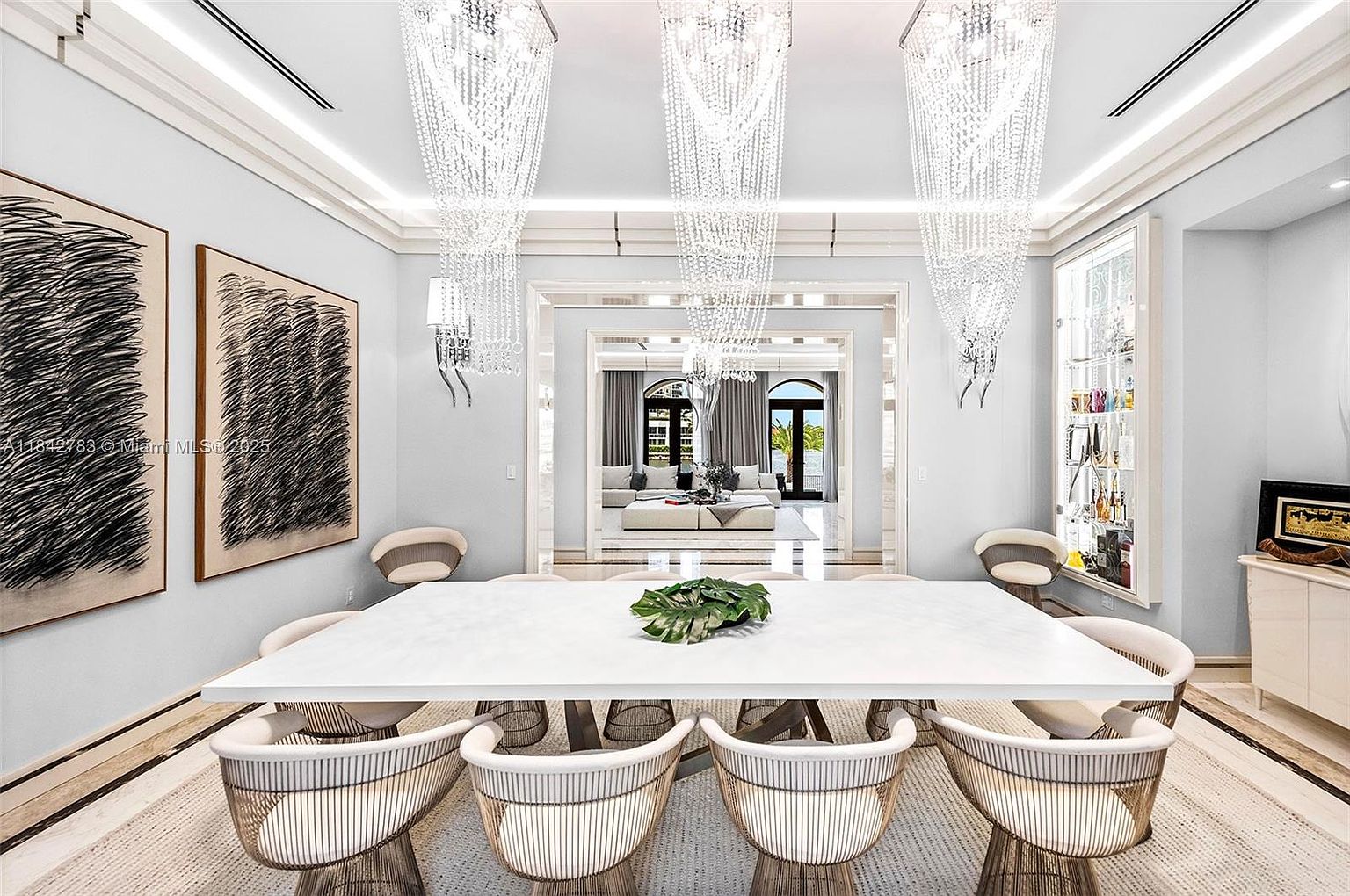 This elegant dining room features a large, rectangular white table surrounded by modern, curved chairs with metallic bases. Three dramatic, tiered crystal chandeliers hang above the table, casting a sophisticated glow over the light-toned space. The room is framed by two large, abstract black-and-white art pieces and offers a clear, symmetrical view through an open doorway into a bright living area beyond.
