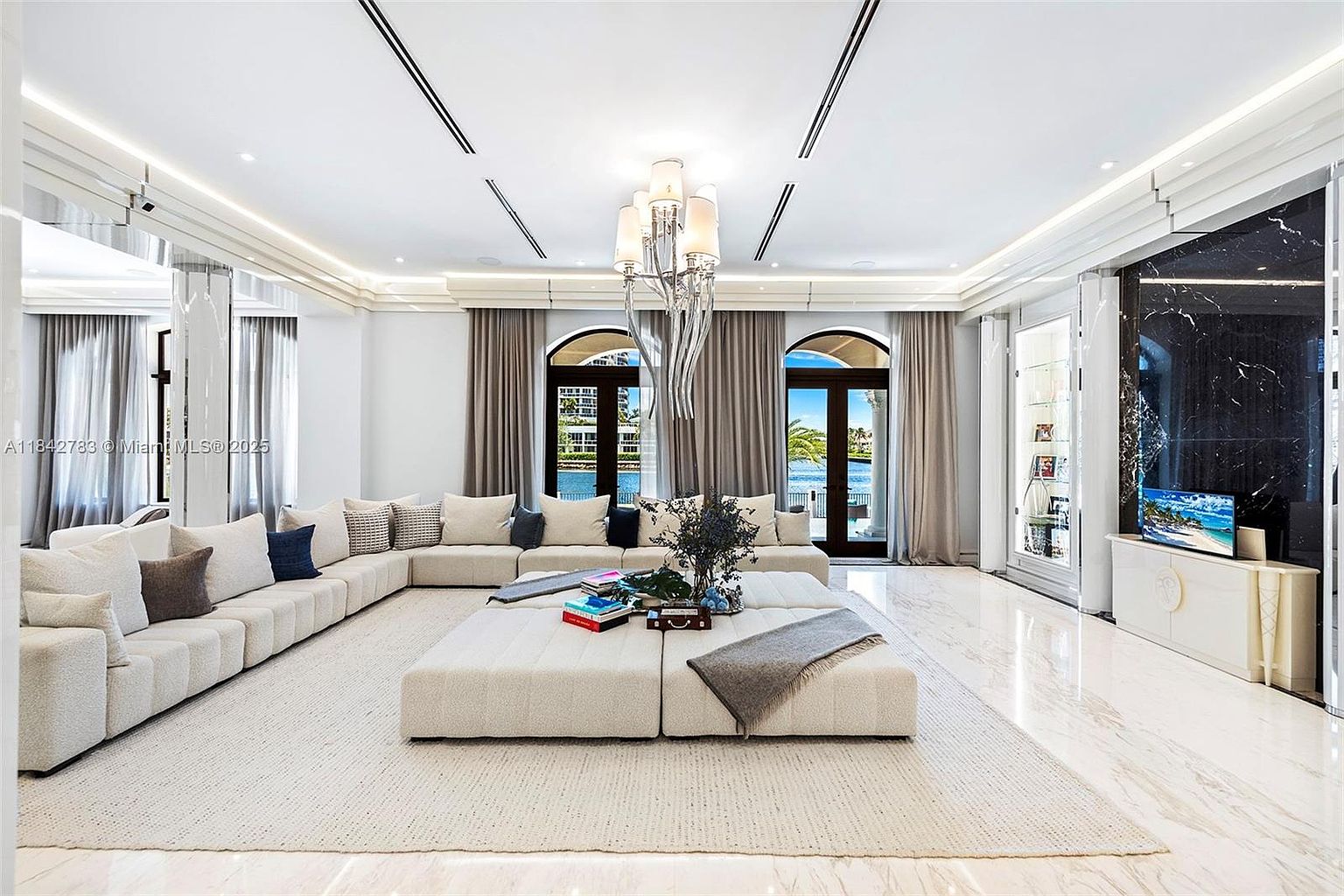 This expansive, high-ceilinged living room features a large, neutral-toned sectional sofa arranged around a central ottoman, creating a spacious seating area. The room is anchored by a polished stone floor and illuminated by a prominent, multi-tiered chandelier, while large glass doors provide a clear view of the waterfront outside. A sleek, dark stone wall serves as a focal point, housing a television and integrated display shelving, contributing to a sophisticated and modern aesthetic.