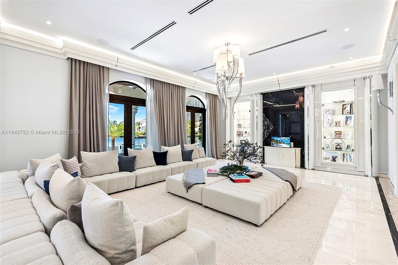 This expansive living room features a neutral color palette with a large, modular sectional sofa arranged around a central ottoman. The space is anchored by a polished stone floor and illuminated by a prominent, multi-tiered crystal chandelier hanging from a recessed ceiling. Large arched windows with floor-to-ceiling curtains provide a view of the water, while a dark, glossy wall panel serves as a dramatic backdrop for a television and display shelving.