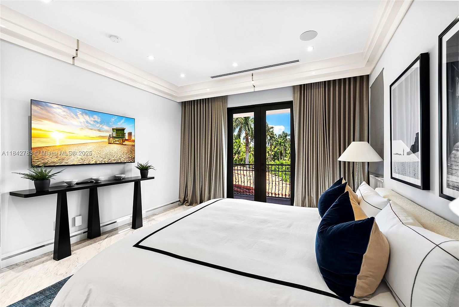 This primary bedroom features a clean, modern aesthetic with a large bed dressed in white linens with black trim and navy accent pillows. A sleek, minimalist console table sits beneath a wall-mounted television, while tall curtains frame a set of double doors that open to a balcony with lush tropical views. The room is illuminated by recessed lighting and a bedside lamp, creating a bright and tranquil atmosphere.