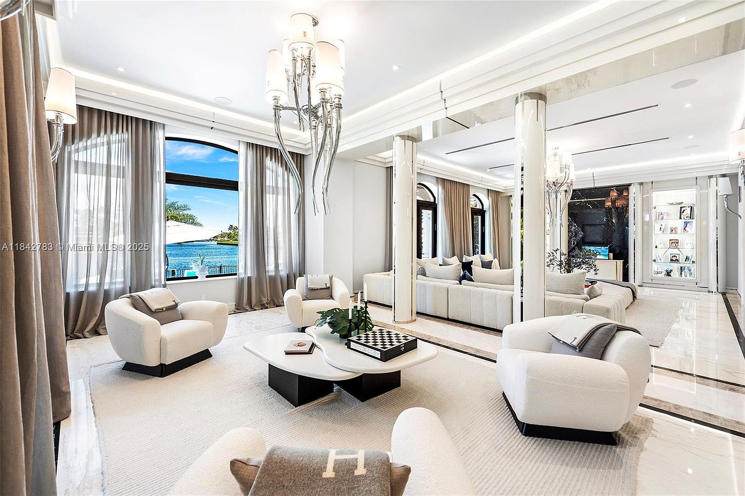 This expansive, high-end living room features a sophisticated neutral color palette with plush, curved white seating arranged around a tiered, modern coffee table. Large windows provide a stunning, unobstructed view of the waterfront, while a dramatic, multi-armed chandelier serves as a focal point above the seating area. The space is defined by polished stone flooring, elegant columns, and a bright, airy atmosphere that emphasizes luxury and open-concept design.
