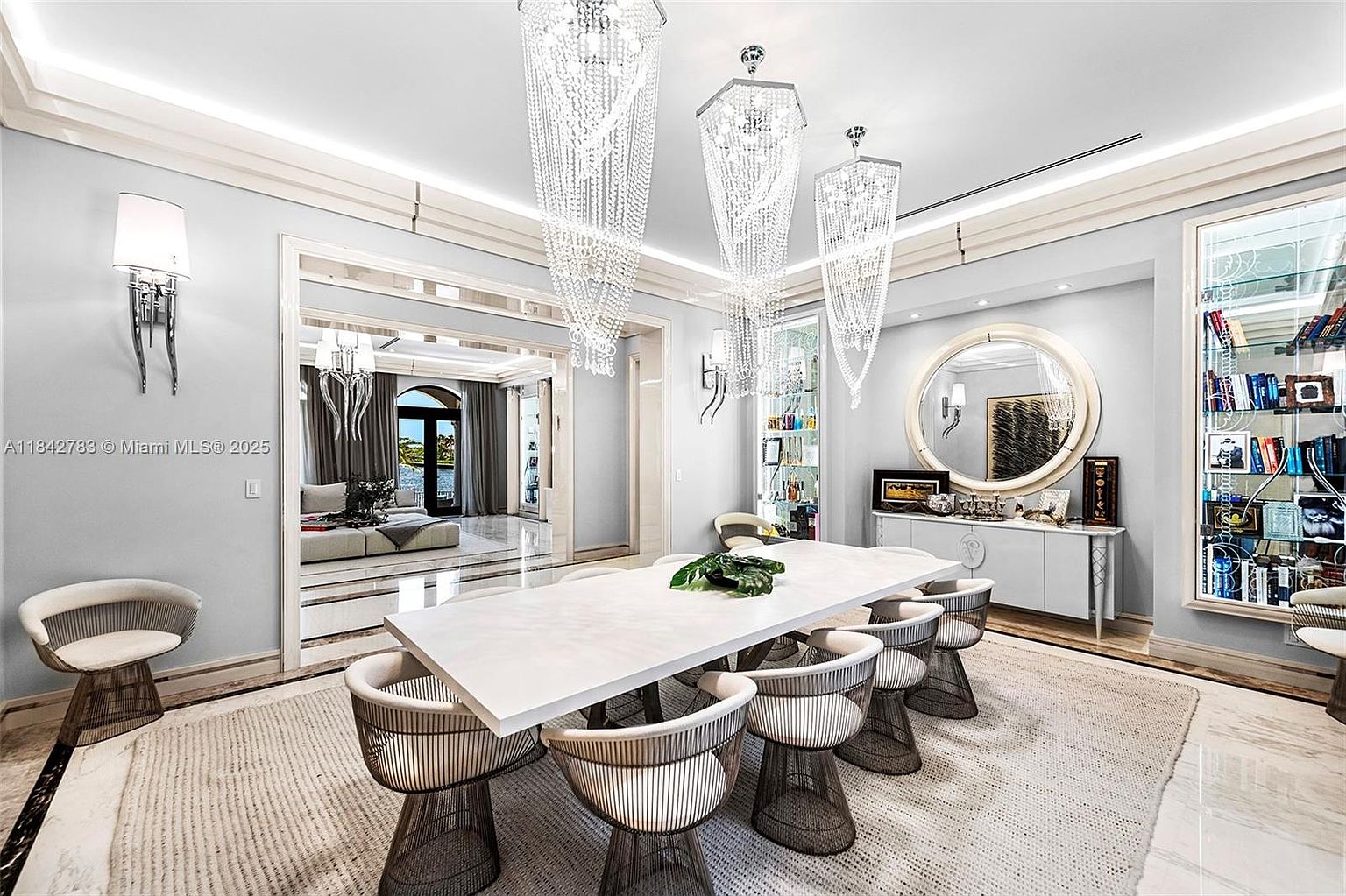 This elegant dining room features a long, light-toned table surrounded by modern, wire-frame chairs set atop a textured area rug. Three dramatic, tiered crystal chandeliers hang from the ceiling, casting a sophisticated glow over the space, which is flanked by built-in glass display cabinets and a circular wall mirror. The room opens into an adjacent living area, creating a seamless flow that highlights the home's spacious and luxurious design.