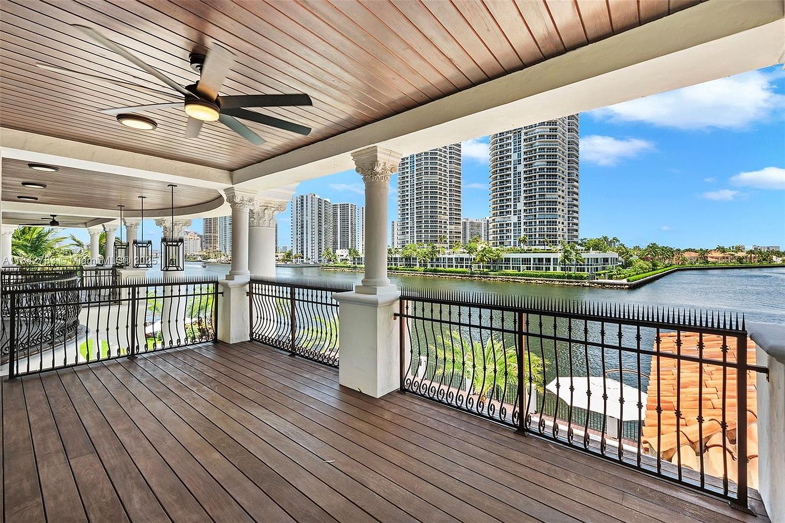 This expansive covered balcony features warm-toned wood plank flooring and a wood-paneled ceiling equipped with a ceiling fan. The space is enclosed by a decorative black metal railing with integrated spikes, offering an unobstructed view of the waterfront and the surrounding high-rise skyline. The perspective captures the transition from the sheltered outdoor living area to the scenic canal and urban landscape beyond.