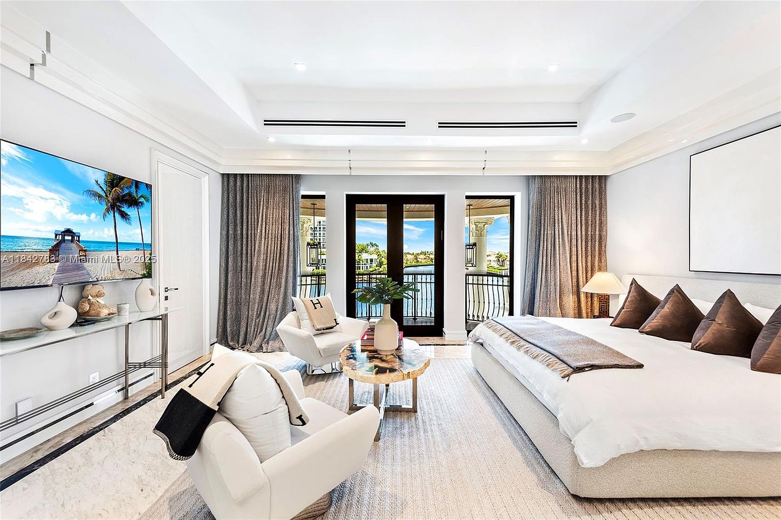This luxurious primary bedroom features a neutral color palette with a large, plush bed and a cozy seating area centered around a round stone-topped coffee table. The room is bathed in natural light from large glass doors that open onto a private balcony overlooking a serene waterfront view. Elegant drapery, recessed lighting, and a wall-mounted television create a sophisticated and tranquil atmosphere.