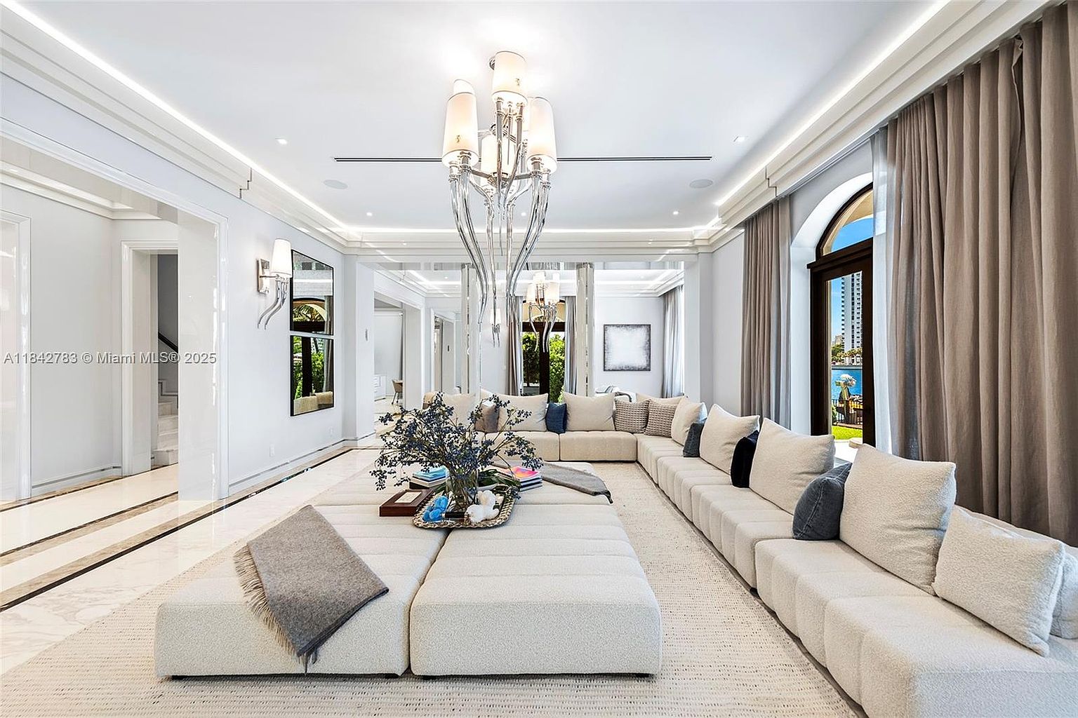 This expansive, light-filled living room features a massive, modular sectional sofa arranged around a central ottoman, creating a luxurious lounge atmosphere. A striking, multi-tiered crystal chandelier serves as the focal point, hanging above the seating area, while large windows with floor-to-ceiling drapery offer views of the outdoors. The space is finished with polished light-toned flooring and recessed lighting, conveying an elegant and sophisticated aesthetic.